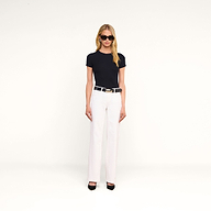 Eine Frau mit langen blonden Haaren, die eine schwarze Sonnenbrille, ein schwarzes kurzärmeliges Oberteil, einen schwarzen Gürtel, eine „Joanne Low-Rise Wide-Leg Jean“ in Weißwaschoptik und schwarze Pumps trägt und vor einem beigen Hintergrund steht.