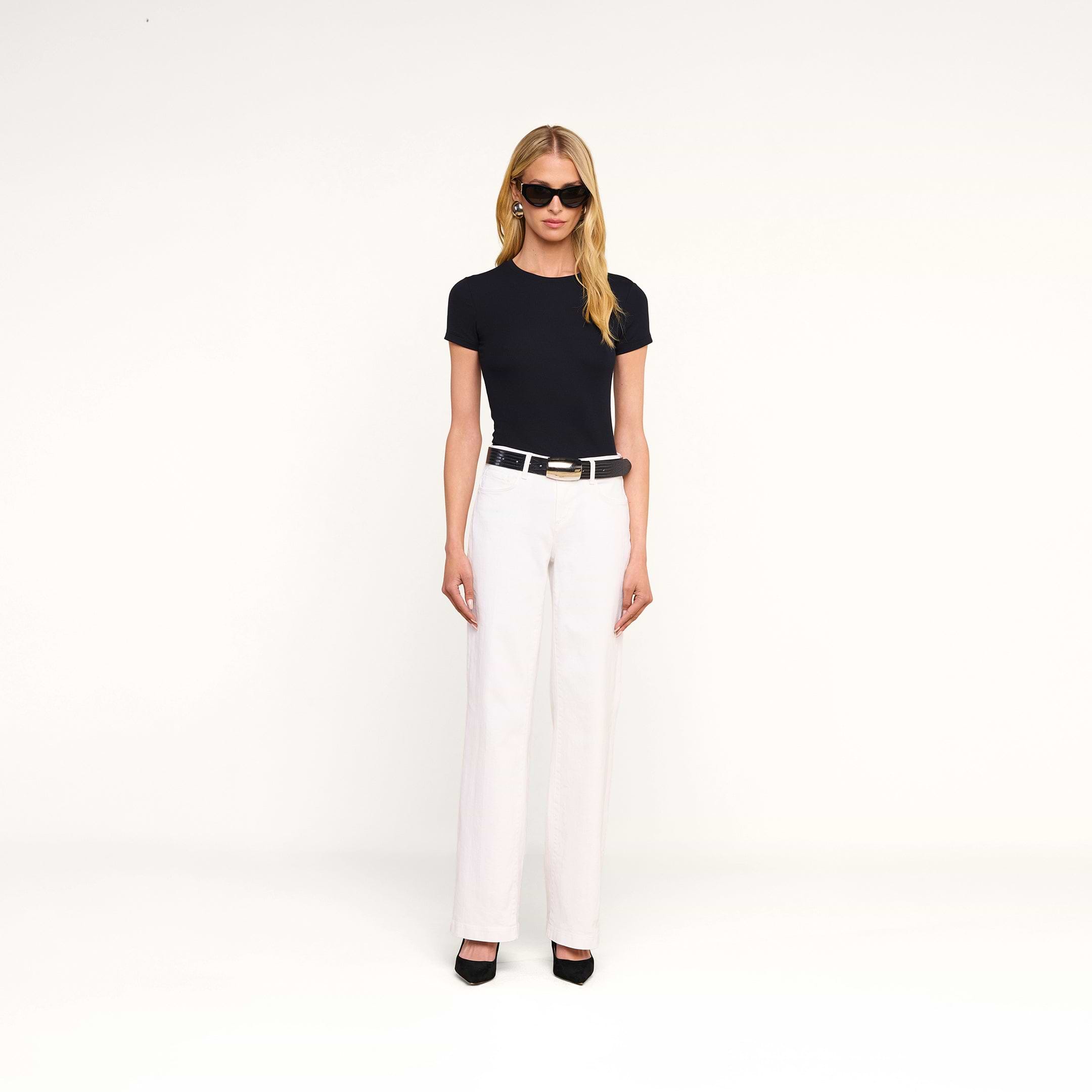 Eine Frau mit langen blonden Haaren, die eine schwarze Sonnenbrille, ein schwarzes kurzärmeliges Oberteil, einen schwarzen Gürtel, eine „Joanne Low-Rise Wide-Leg Jean“ in Weißwaschoptik und schwarze Pumps trägt und vor einem beigen Hintergrund steht.