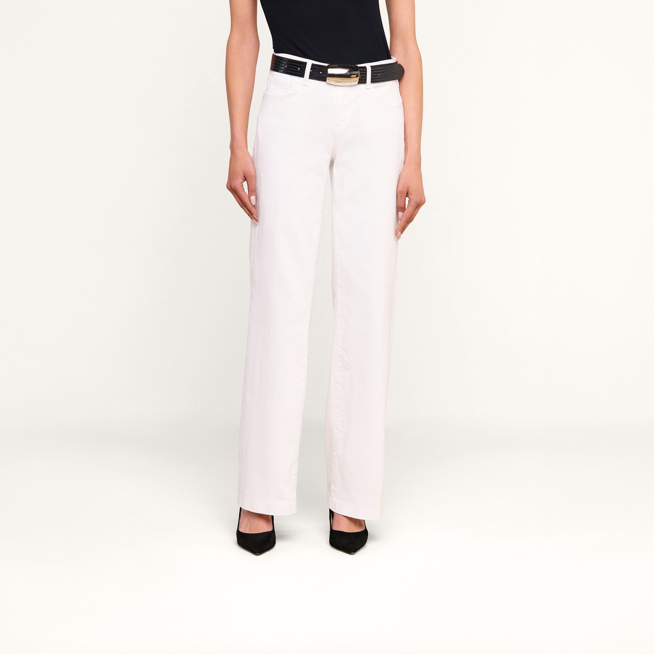 Eine Person, die eine weiße „Joanne Low-Rise Wide-Leg Jeans“ trägt, dazu einen schwarzen Gürtel mit goldener Schnalle, ein schwarzes Oberteil und schwarze Schuhe mit hohen Absätzen, steht vor einem beigen Hintergrund.