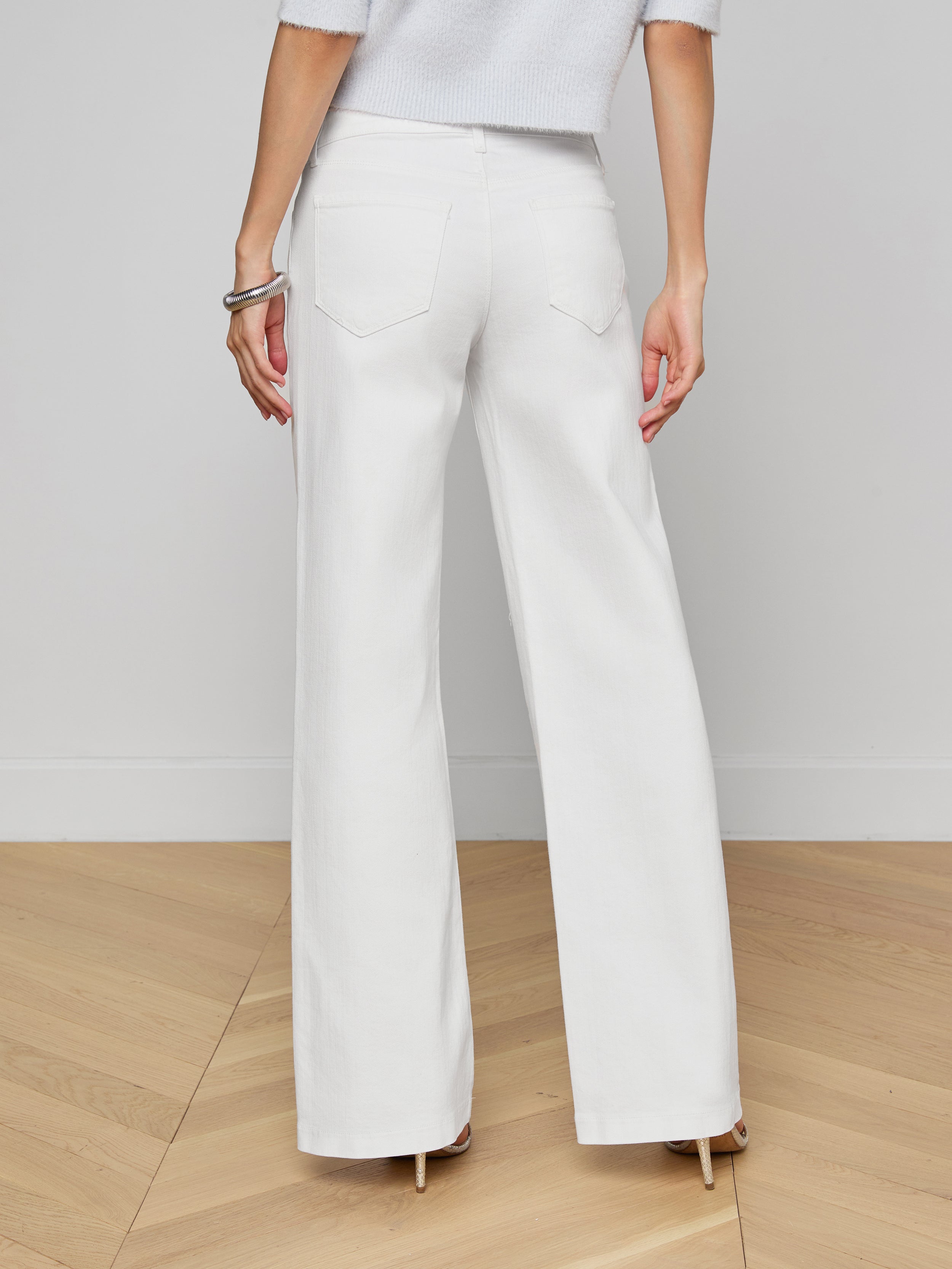 Person standing facing away, wearing Joanne Low-Rise Wide-Leg Jeans with a black belt and black high heels, against a plain white background. Only the lower half of the body is visible.