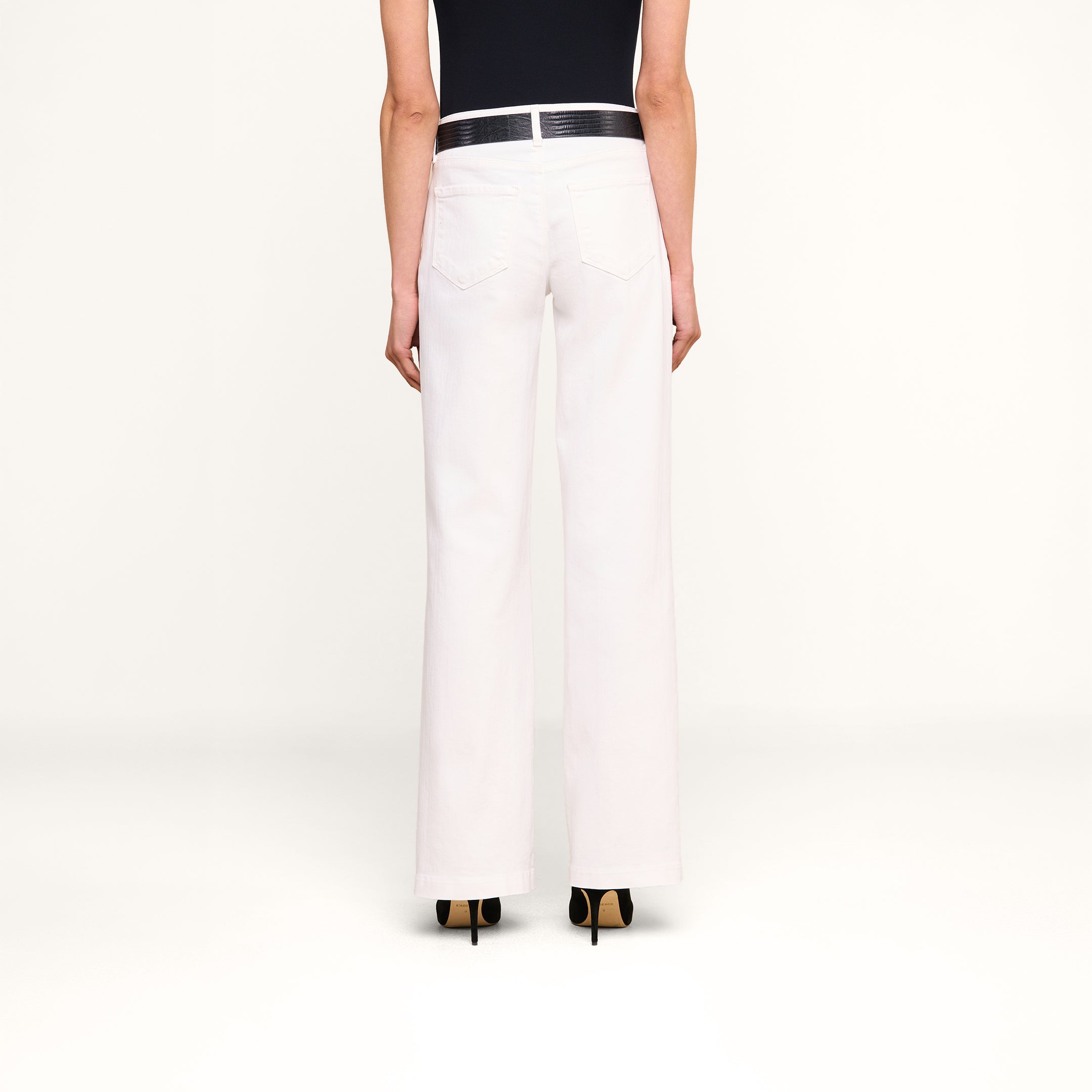 Eine Person ist von hinten vor einem beigen Hintergrund zu sehen. Sie trägt eine weiße „Joanne Low-Rise Wide-Leg Jeans“, einen schwarzen Gürtel und schwarze Schuhe mit Absatz.