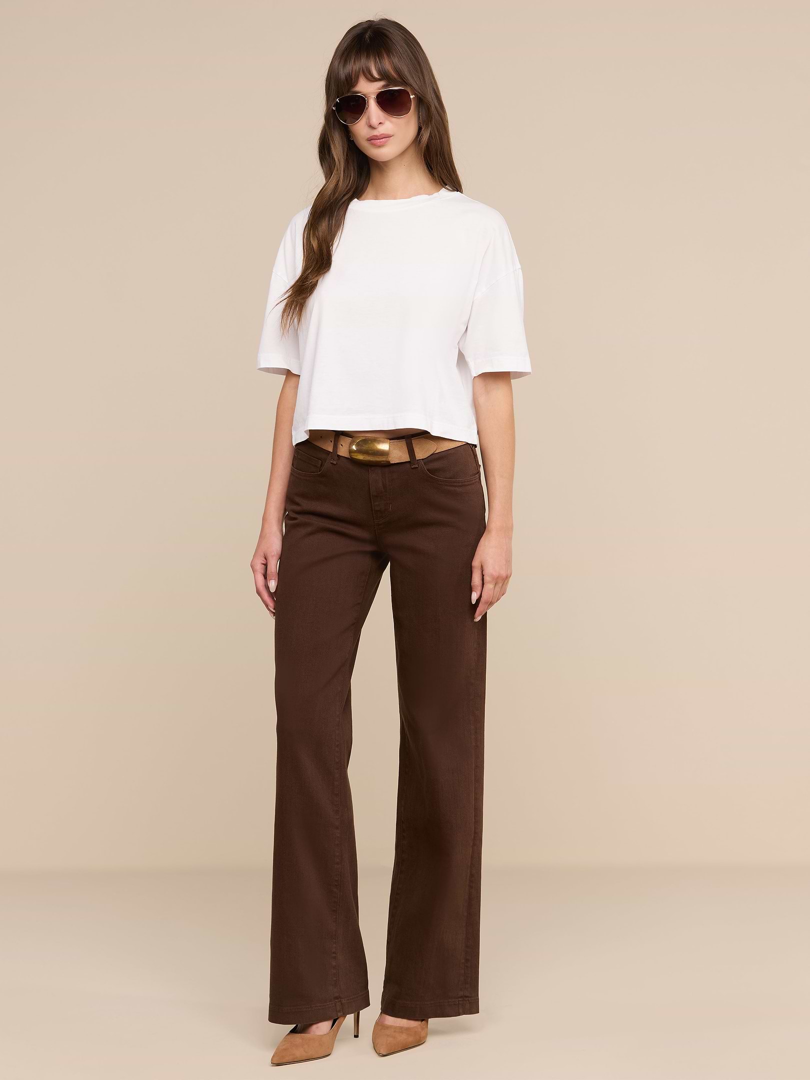 Une femme se tient debout devant un fond beige ; elle porte des lunettes de soleil, un haut court blanc à manches courtes, des talons marron et le jean « Joanne Low-Rise Wide-Leg » agrémenté dune ceinture en denim extensible.