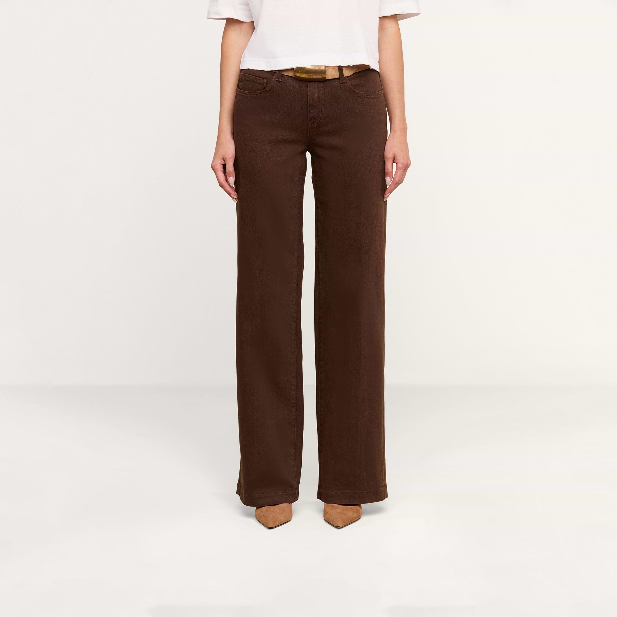 Une personne porte un haut blanc à manches courtes, un jean « Joanne » taille basse à jambes larges en denim stretch marron, ainsi que des chaussures à bout pointu de couleur beige clair, debout devant un fond beige. La photo est cadrée au niveau des épaules.