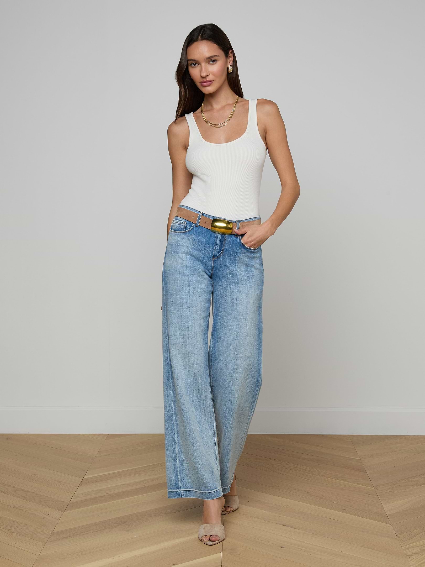 A woman wearing a white tank top, beige heels, and a belt stands against a plain white background in light blue Joanne Low-Rise Wide-Leg Jeans.