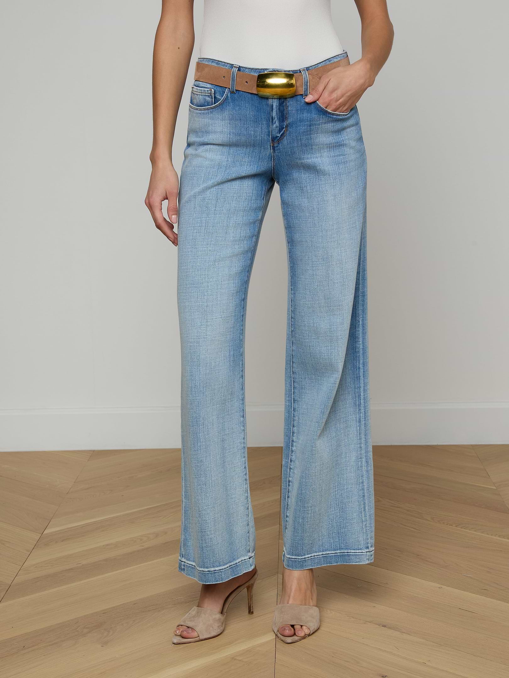 A woman wears the Joanne Low-Rise Wide-Leg Jean in light blue, paired with a brown belt, white sleeveless top, and beige heeled sandals. She stands against a plain light background with only the lower half of her body visible.