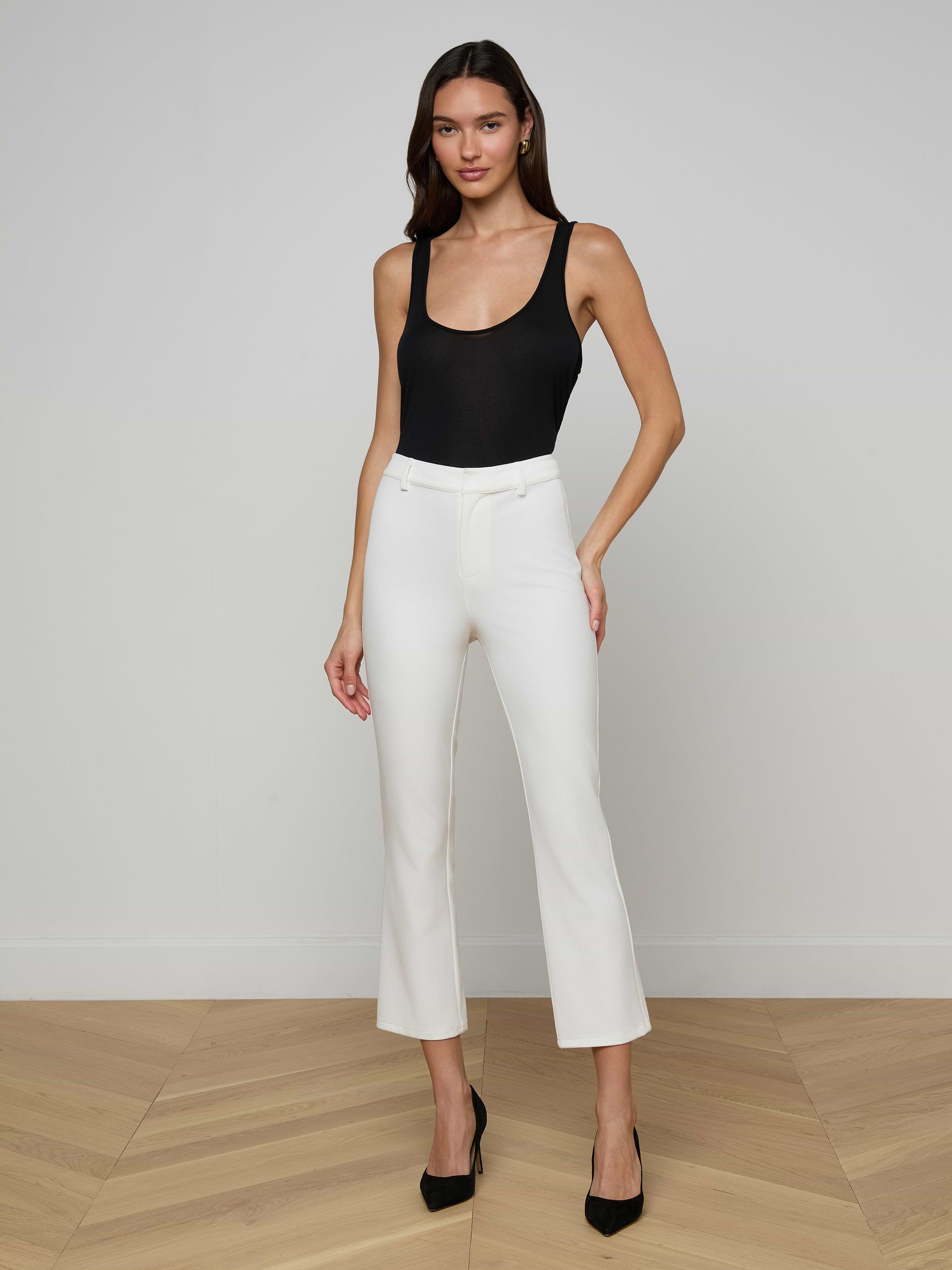 A woman stands against a plain white background, posing with one hand on her hip while wearing a black sleeveless top, black heels, and white Ketra Bootcut Trousers crafted from stretch suit fabric.