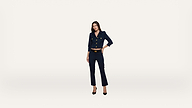 A woman in a navy blue suit with gold buttons stands confidently against a plain white background, her left hand on her hip and black heels on her feet.