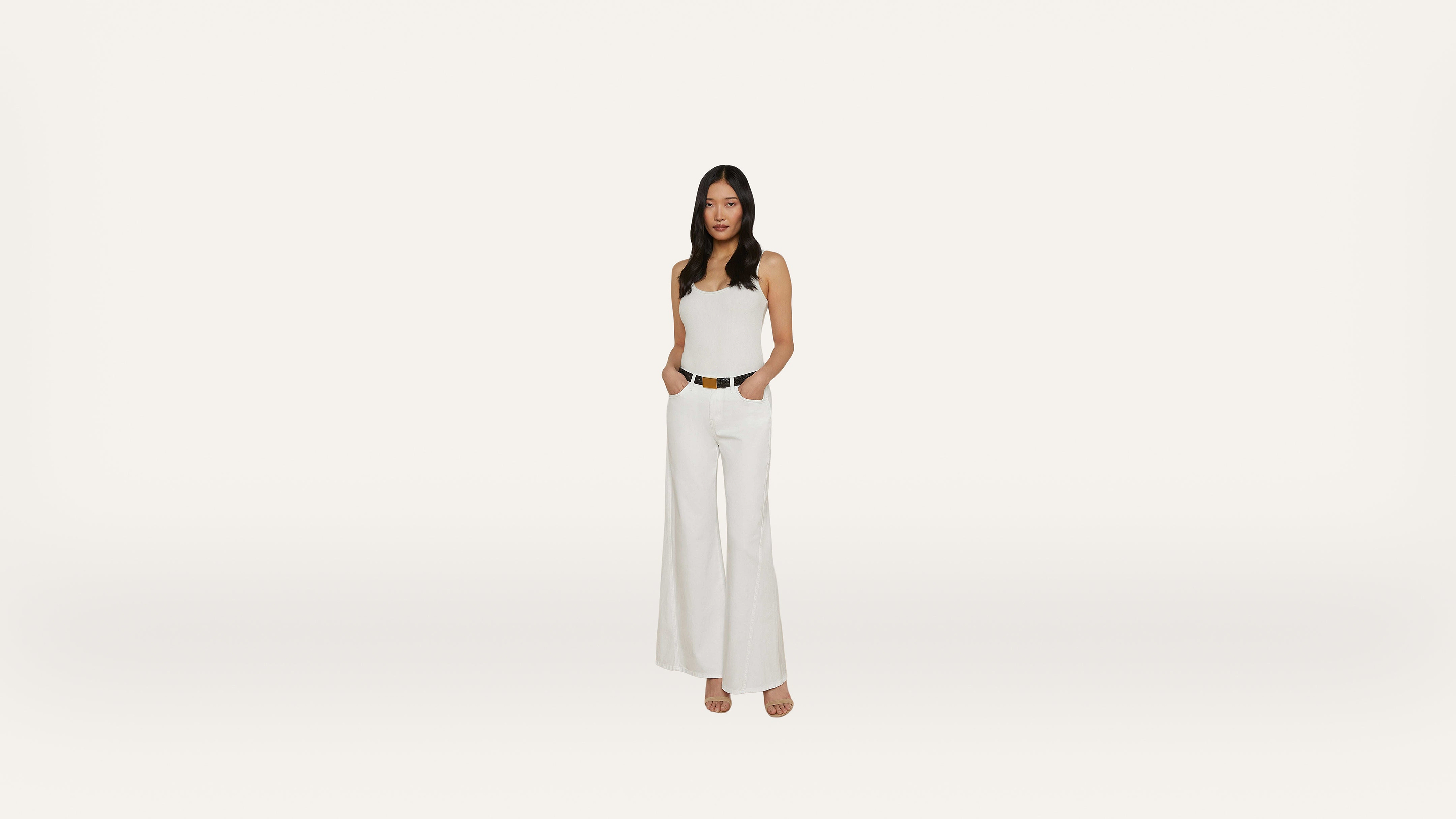 Woman wearing a white sleeveless top and wide-leg white pants, standing with hands in pockets against a plain white background.