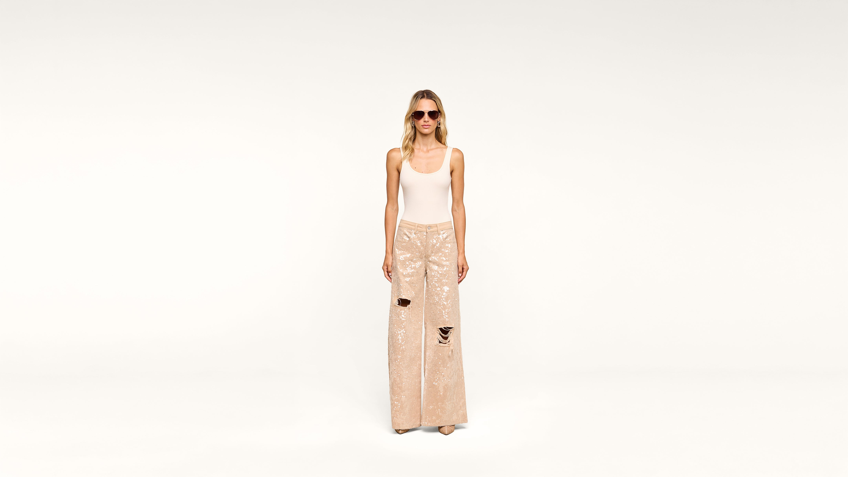 Woman wearing sunglasses, a white tank top, and wide-legged, shiny beige sequin pants with rips, standing against a plain white background.