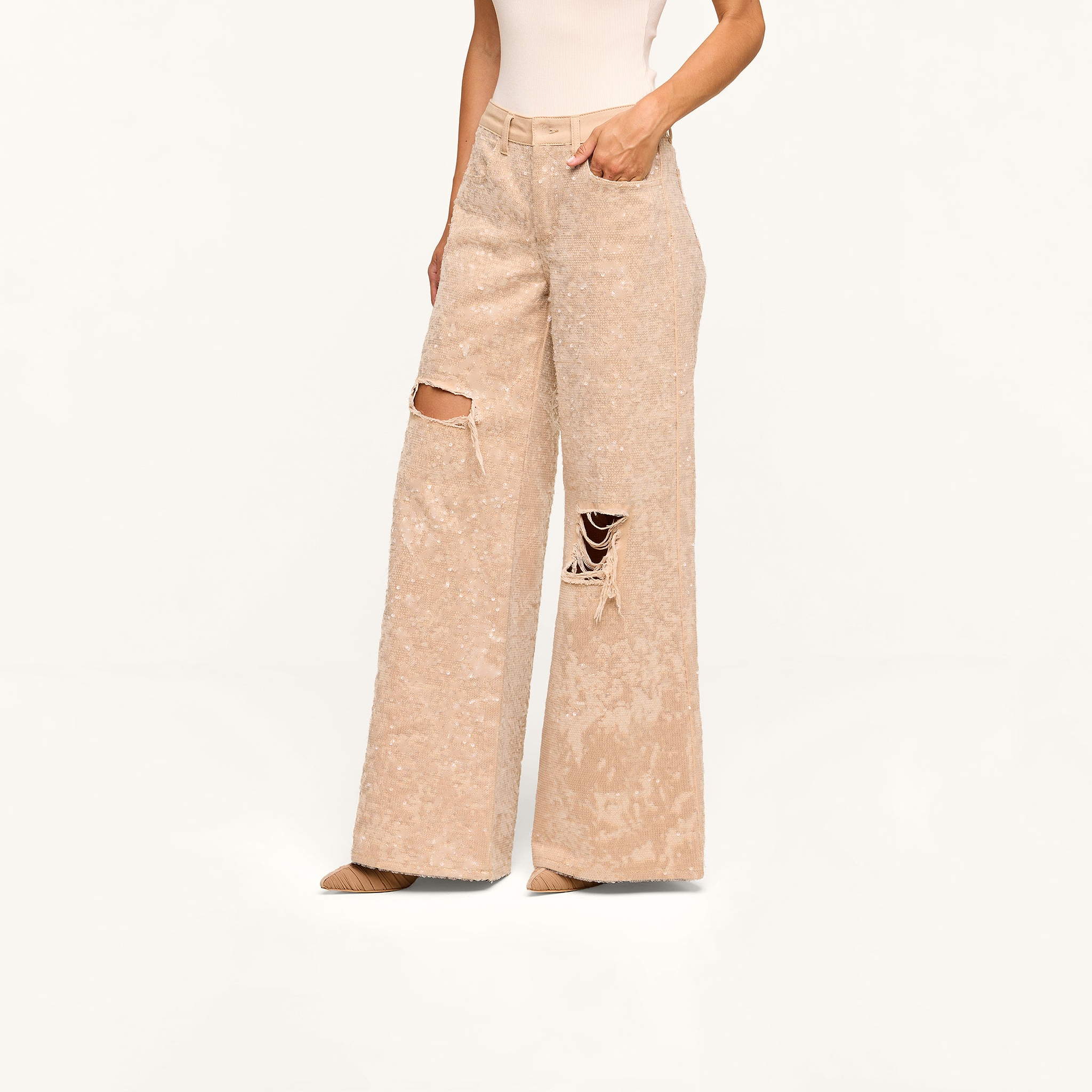 Wearing the Quincy Embellished Wide-Leg Jean with ripped knees, a beige sleeveless top, and beige shoes, this person stands against a plain white background.