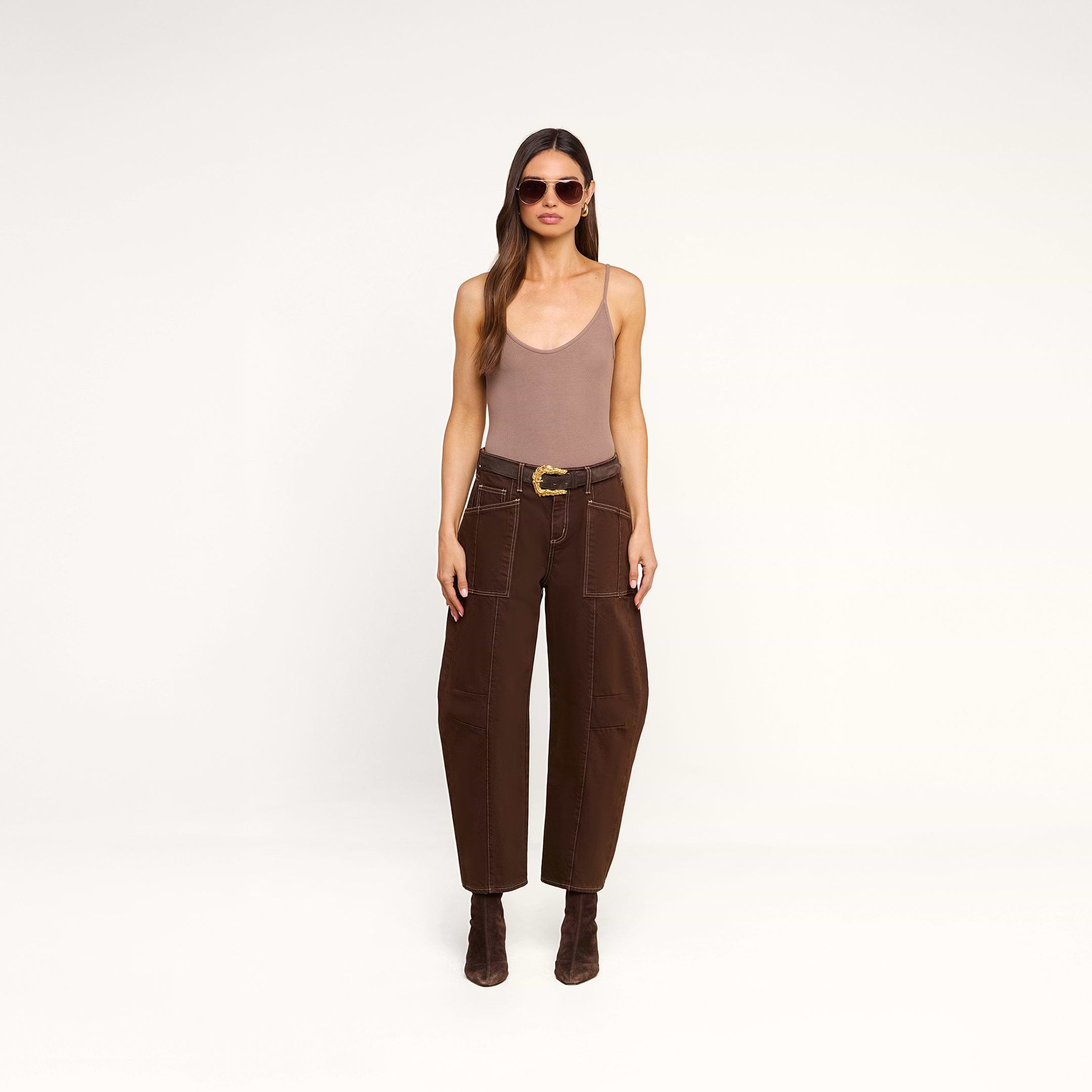 A woman wearing sunglasses, a beige tank top, the Cody Cropped Barrel-Leg Jean in brown with large pockets, a wide belt, and brown boots stands against a plain white background.