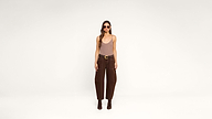 A woman wearing sunglasses, a beige tank top, brown high-waisted pants, and ankle boots stands against a plain white background.