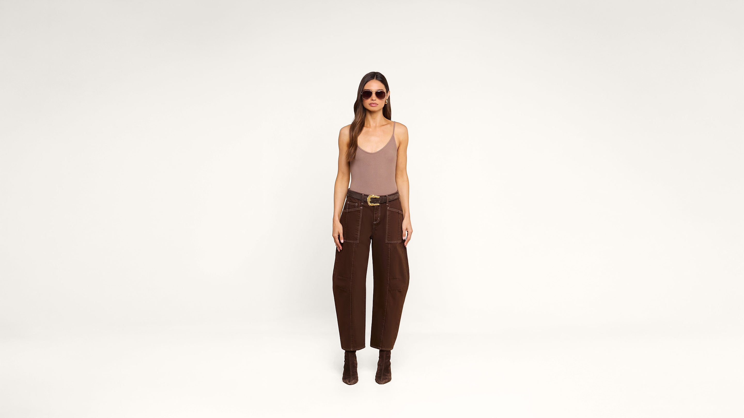 A woman wearing sunglasses, a beige tank top, brown high-waisted pants, and ankle boots stands against a plain white background.