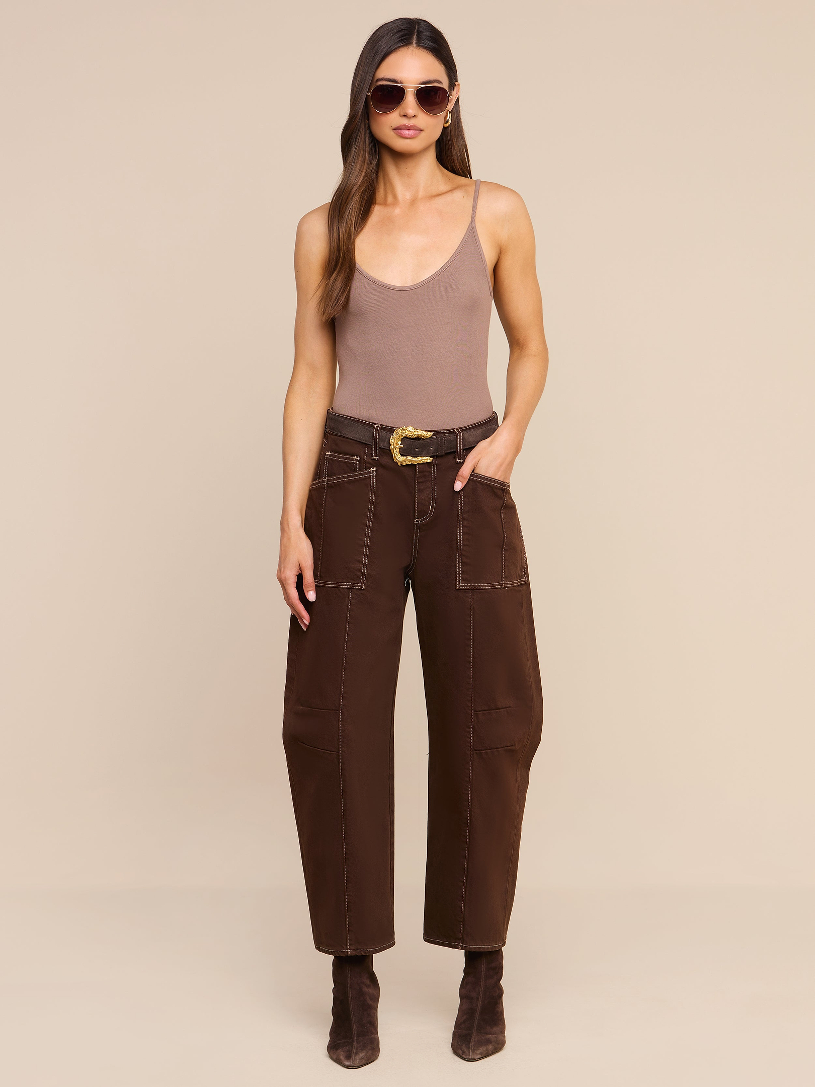 A woman stands against a plain white background wearing sunglasses, a taupe tank top, the Cody Cropped Barrel-Leg Jean in high rise dark brown with patch pockets and a belt, and dark brown boots.