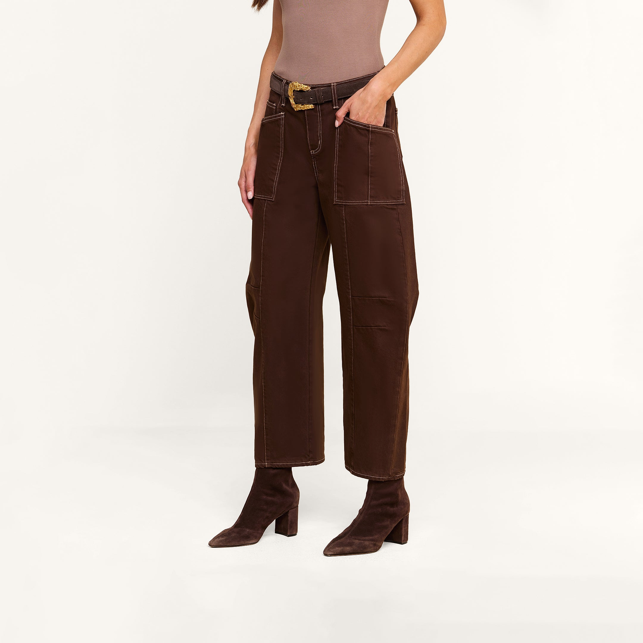 A woman stands against a plain white background wearing a taupe top paired with the Cody Cropped Barrel-Leg Jean, featuring high-rise, wide-leg brown pants with large pockets and a matching belt, and completes the look with brown heeled boots.