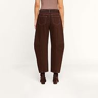 A person stands facing away, wearing the Cody Cropped Barrel-Leg Jean in high-rise, wide-leg brown non-stretch denim with visible white stitching, paired with brown heeled boots. The background is plain and light-colored.