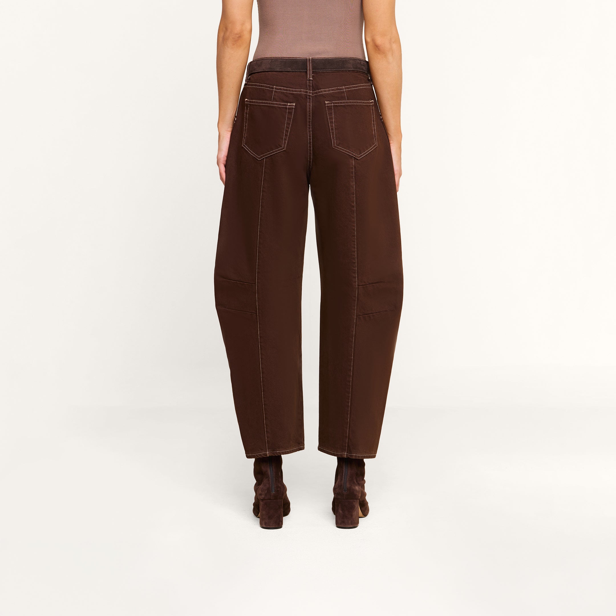 A person stands facing away, wearing the Cody Cropped Barrel-Leg Jean in high-rise, wide-leg brown non-stretch denim with visible white stitching, paired with brown heeled boots. The background is plain and light-colored.