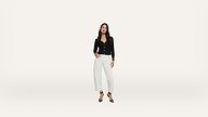 A woman stands against a plain white background, wearing a black long-sleeve top, white high-waisted pants, and black heeled sandals. She has long dark hair and a relaxed pose.
