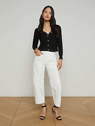 A woman stands against a plain white background, wearing the Cody Barrel-Leg Jean in high-rise white denim, paired with a black long-sleeve top and black open-toe heeled sandals. Her hair is long and loose, and she poses with one hand on her hip.