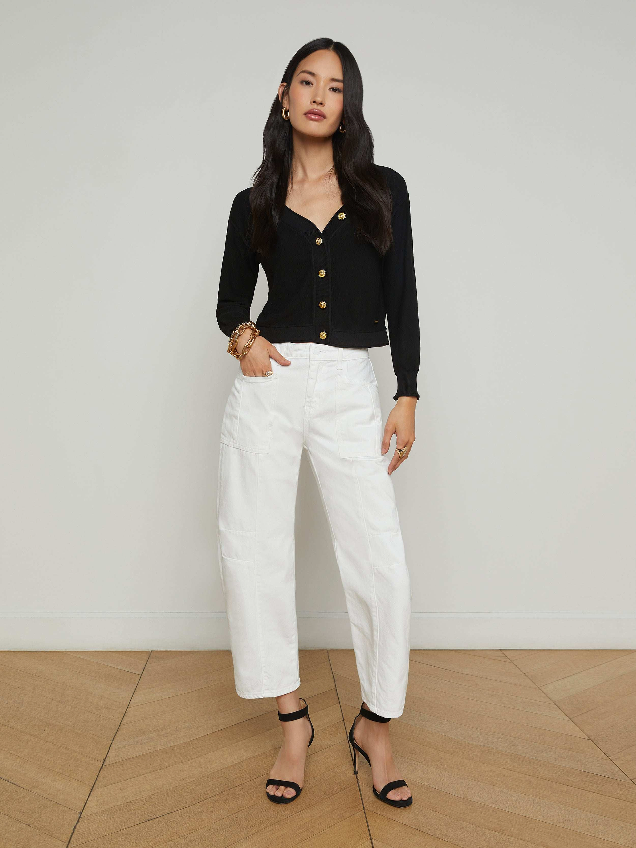 A woman stands against a plain white background, wearing the Cody Barrel-Leg Jean in high-rise white denim, paired with a black long-sleeve top and black open-toe heeled sandals. Her hair is long and loose, and she poses with one hand on her hip.