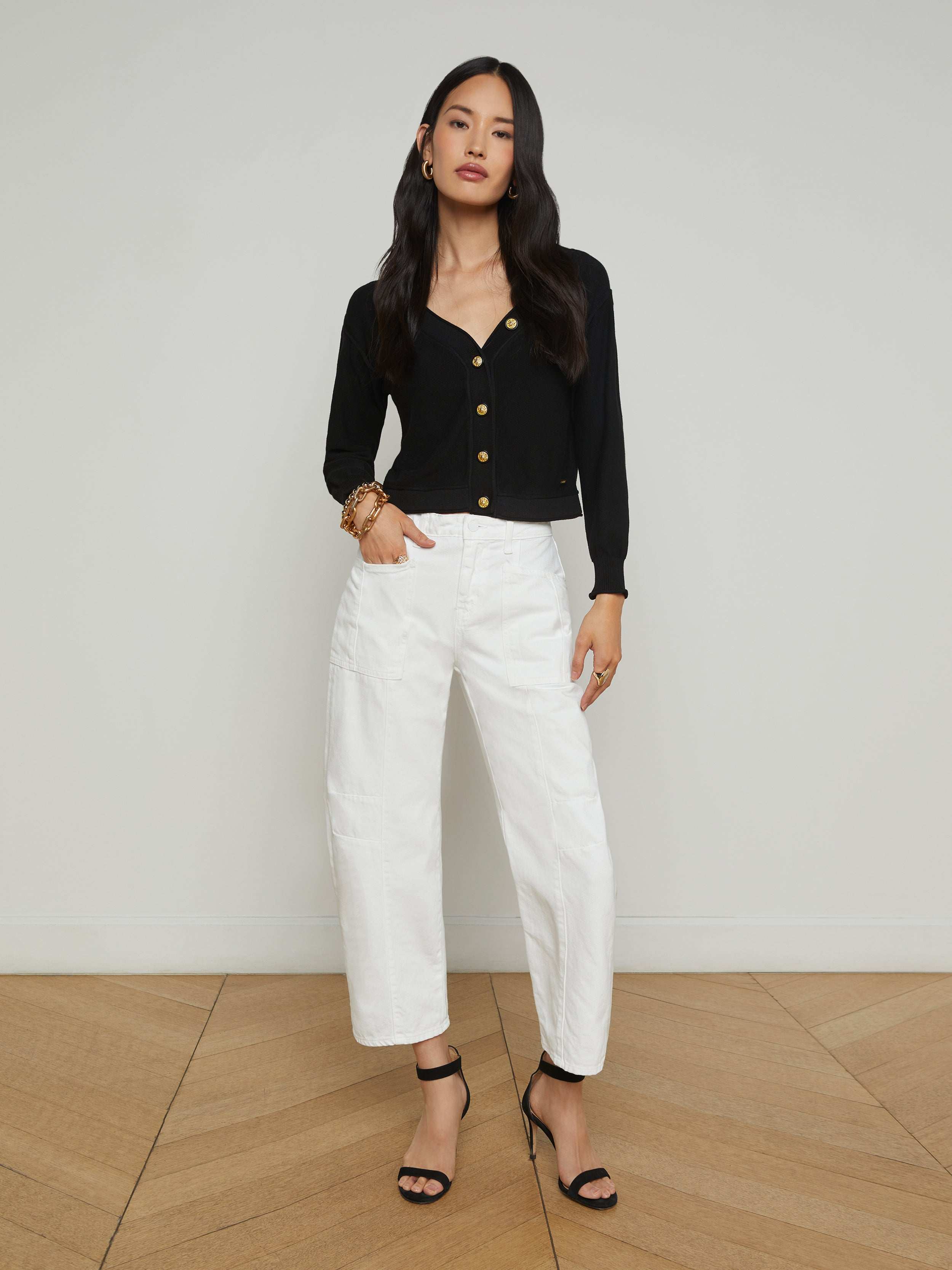 A woman stands against a plain white background, wearing the Cody Barrel-Leg Jean in high-rise white denim, paired with a black long-sleeve top and black open-toe heeled sandals. Her hair is long and loose, and she poses with one hand on her hip.