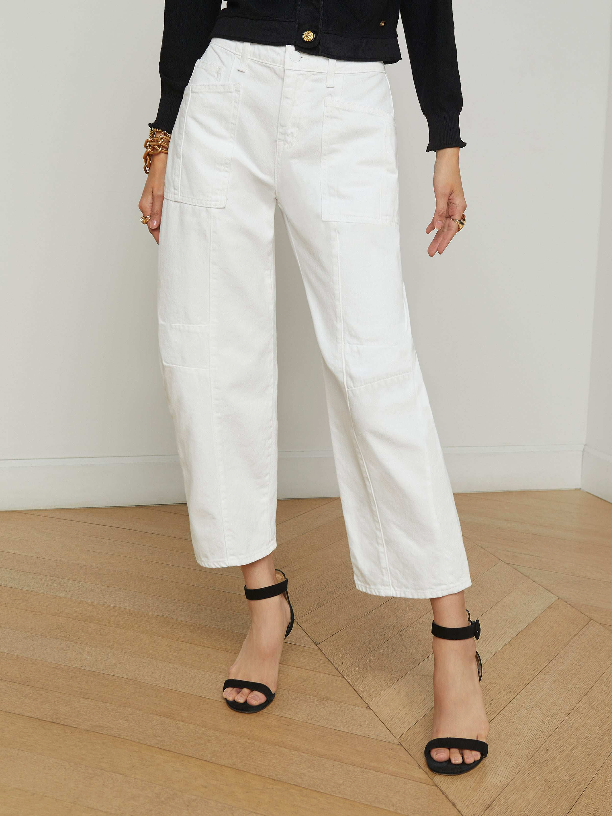 A person wearing the Cody Barrel-Leg Jean in a high-rise white denim style, paired with black heeled sandals, a black long-sleeve top, and bracelets, stands against a plain light background.