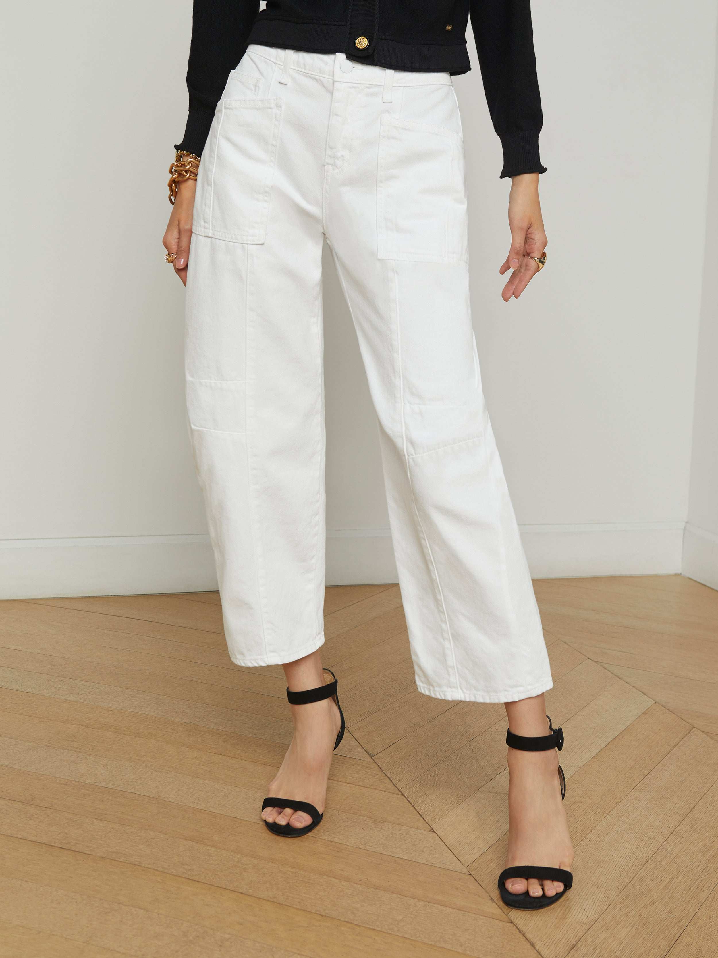 A person wearing the Cody Barrel-Leg Jean in a high-rise white denim style, paired with black heeled sandals, a black long-sleeve top, and bracelets, stands against a plain light background.