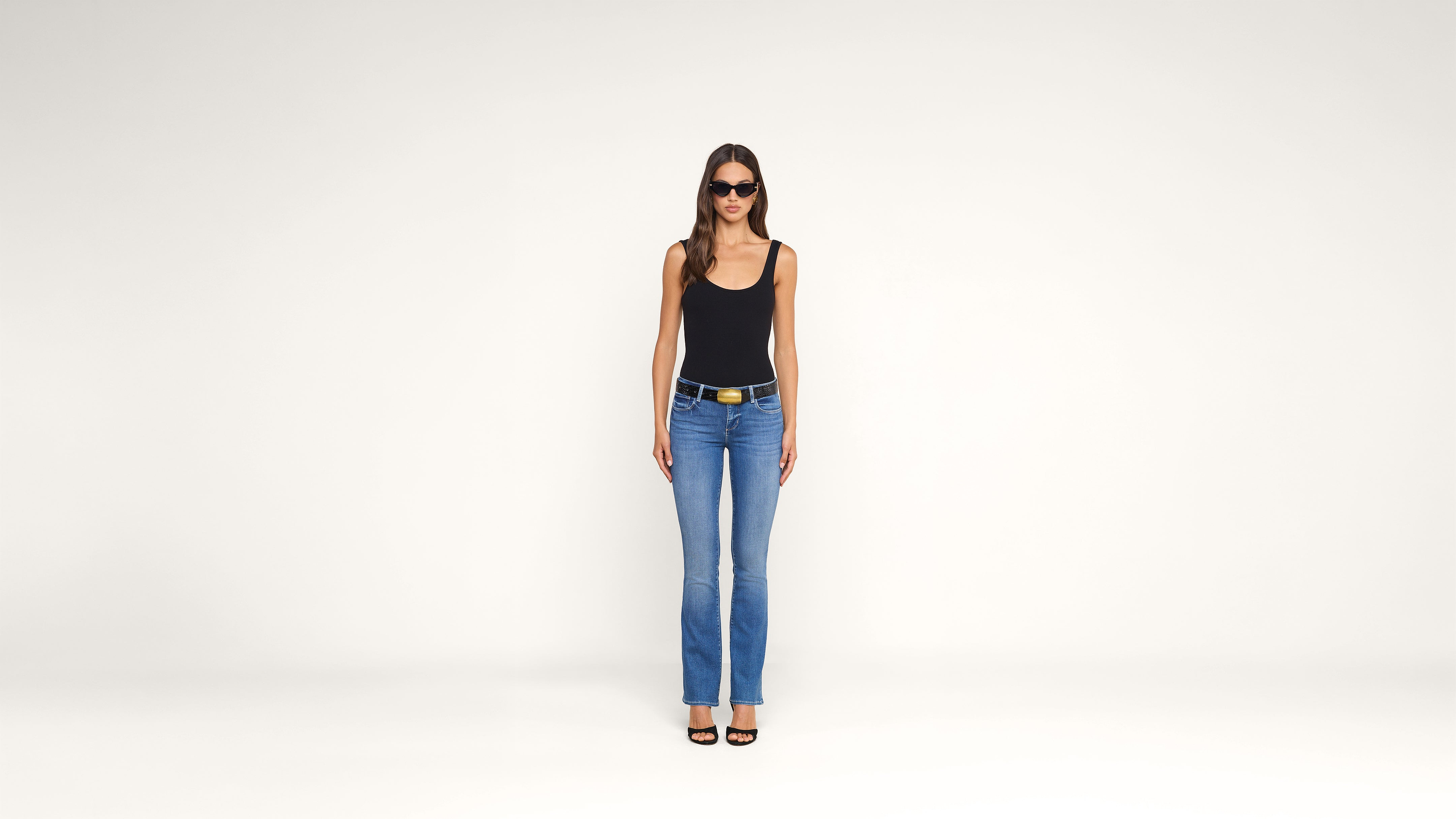 A woman wearing sunglasses, a black tank top, blue jeans, and black sandals stands facing forward against a plain white background.