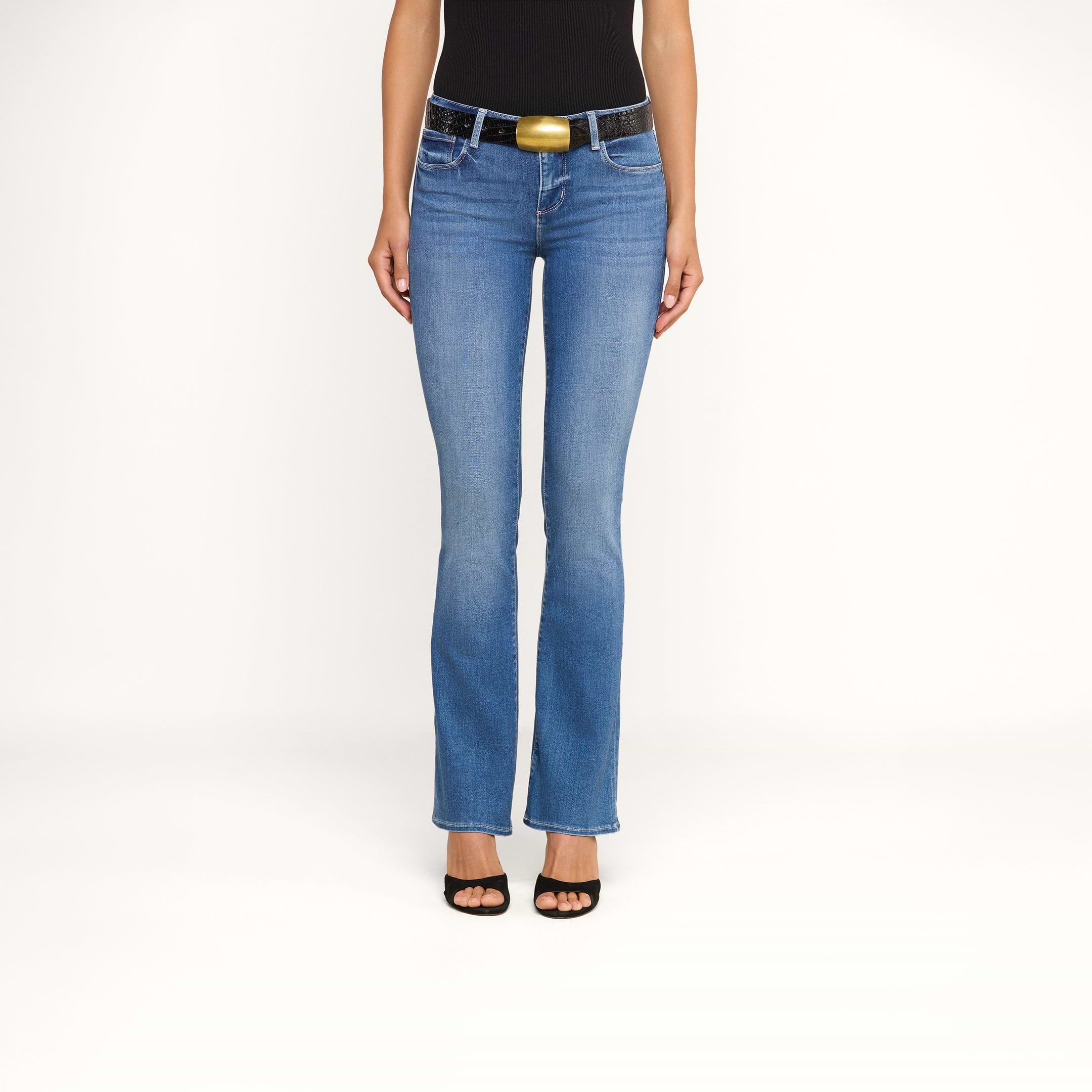 Indossando i jeans Cara a vita bassa e taglio bootcut, un top nero senza maniche, una cintura nera con una grande fibbia dorata e sandali neri con tacco, una persona è in piedi su uno sfondo beige. Il suo volto non è visibile.