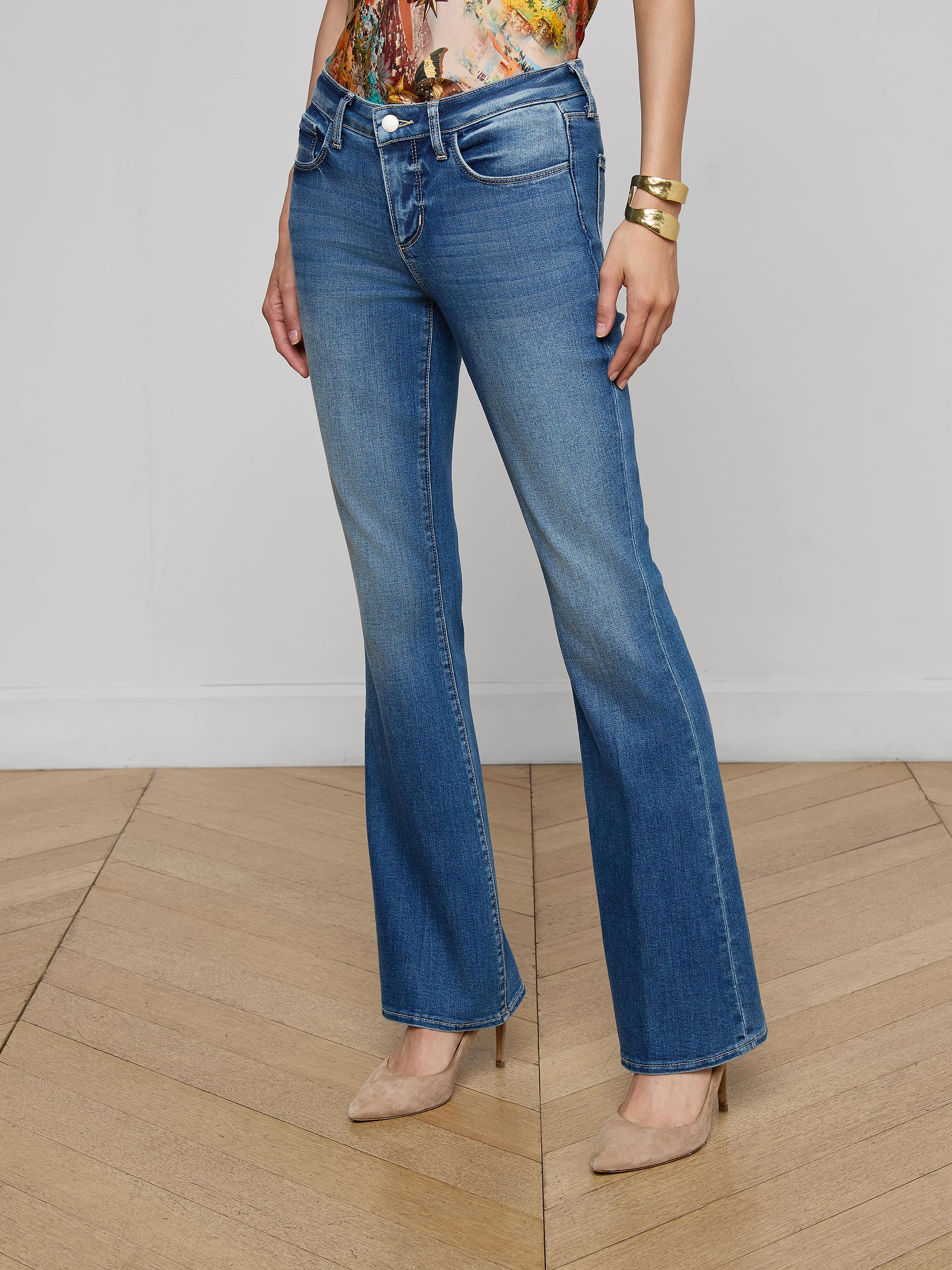 A woman stands against a plain white background wearing the Cara Low-Rise Bootcut Jean in light blue stretch denim, paired with a black belt featuring a large buckle, a black top, and black high-heeled sandals.