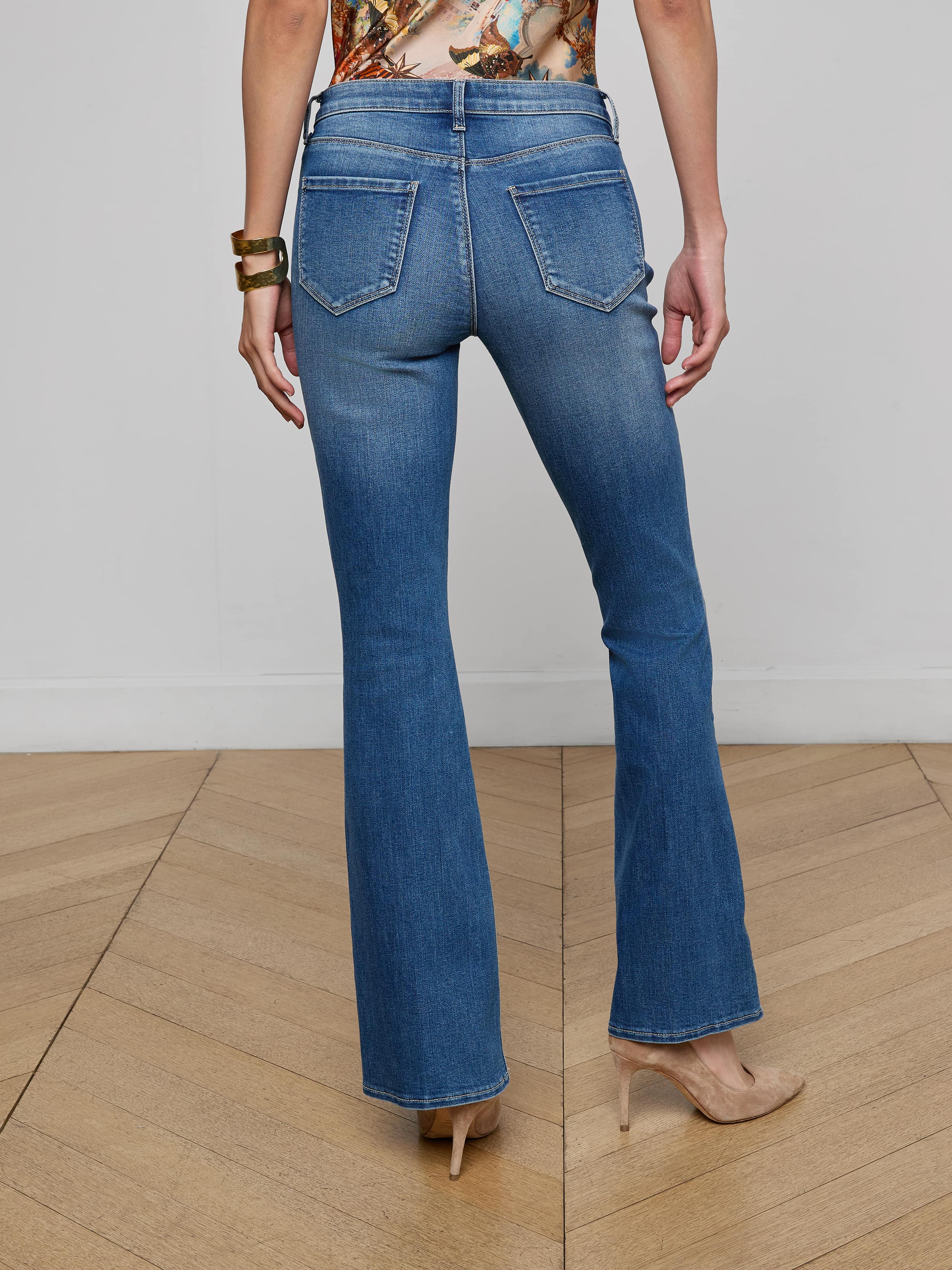 A woman wearing the Cara Low-Rise Bootcut Jean in blue stretch denim, paired with a black belt, black top, and black heeled sandals, is standing with her back to the camera against a plain white background.