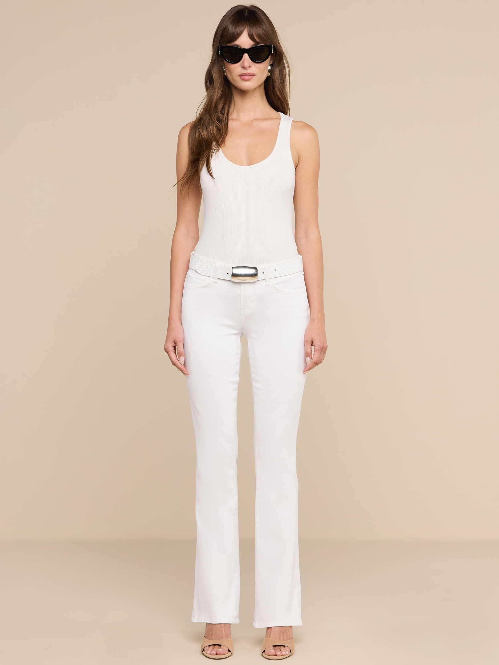 Une femme se tient debout, face à lobjectif, devant un fond beige uni. Elle porte des lunettes de soleil, un haut blanc sans manches, des escarpins ouverts à lavant et le jean « Cara Low-Rise Bootcut » en denim stretch.