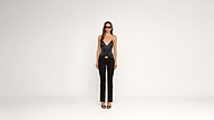 A woman wearing sunglasses, a black sleeveless top, black pants with a belt, and black heels stands against a plain white background.