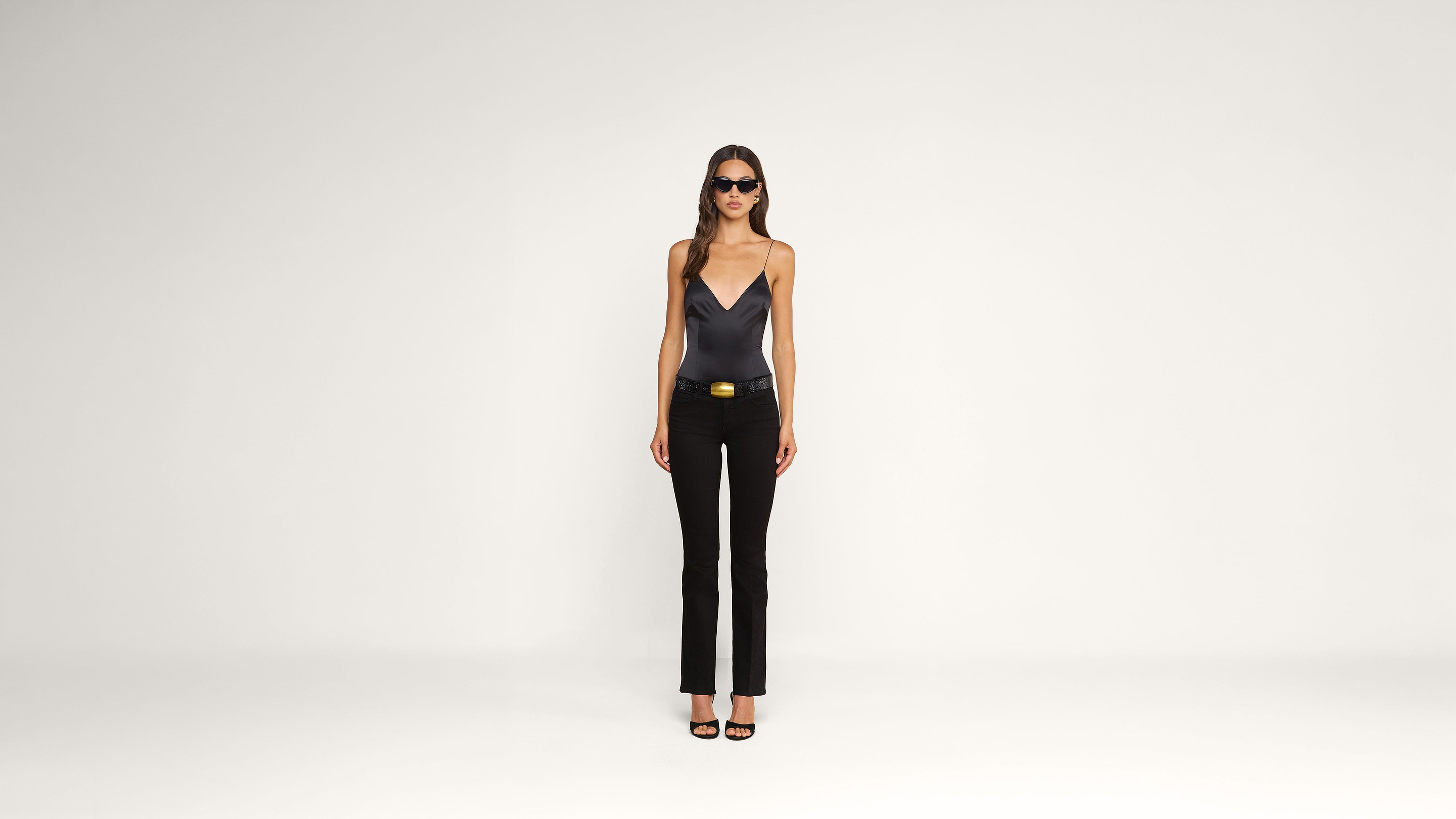 A woman wearing sunglasses, a black sleeveless top, black pants with a belt, and black heels stands against a plain white background.