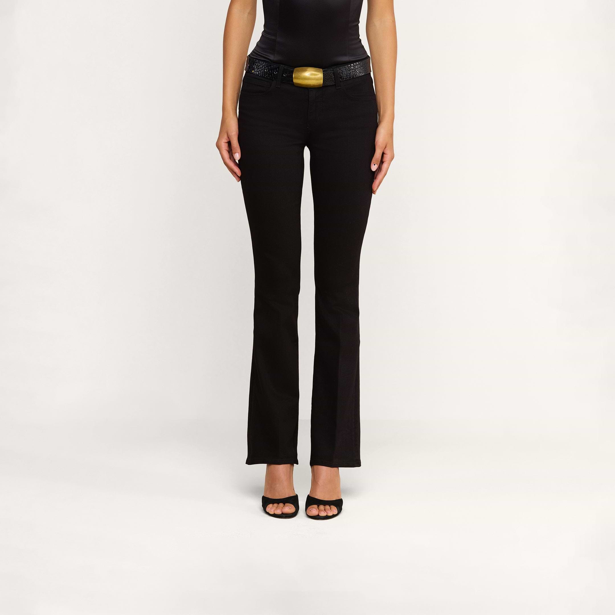 Une personne vêtue dun haut noir sans manches, dun jean bootcut taille basse « Cara » agrémenté dune large ceinture texturée dotée dune grosse boucle dorée, et descarpins noirs à bout ouvert, se tient debout devant un fond beige uni.
