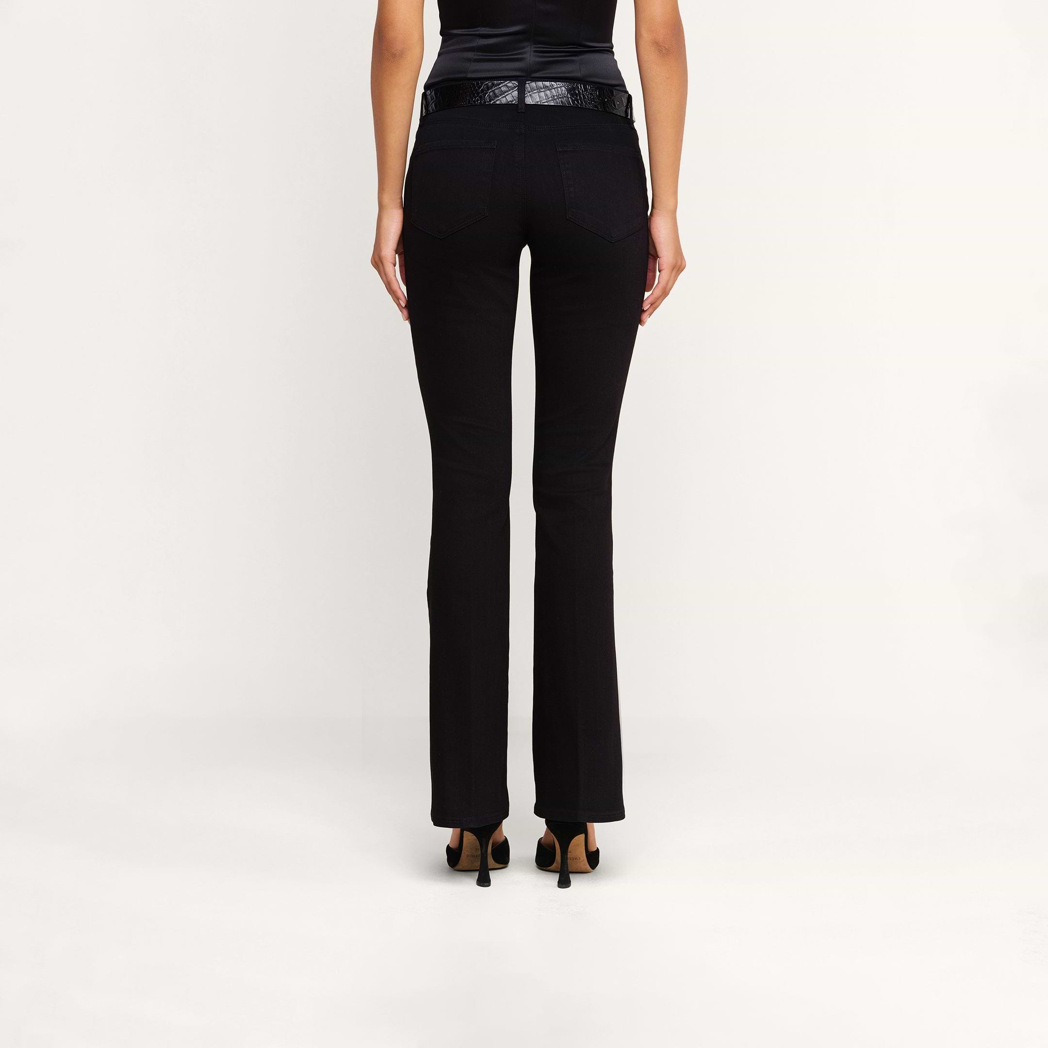 A person stands facing away, wearing the Cara Low-Rise Bootcut Jean in black stretch denim, paired with a black belt, a sleeveless black top, and black heeled shoes against a plain white background.