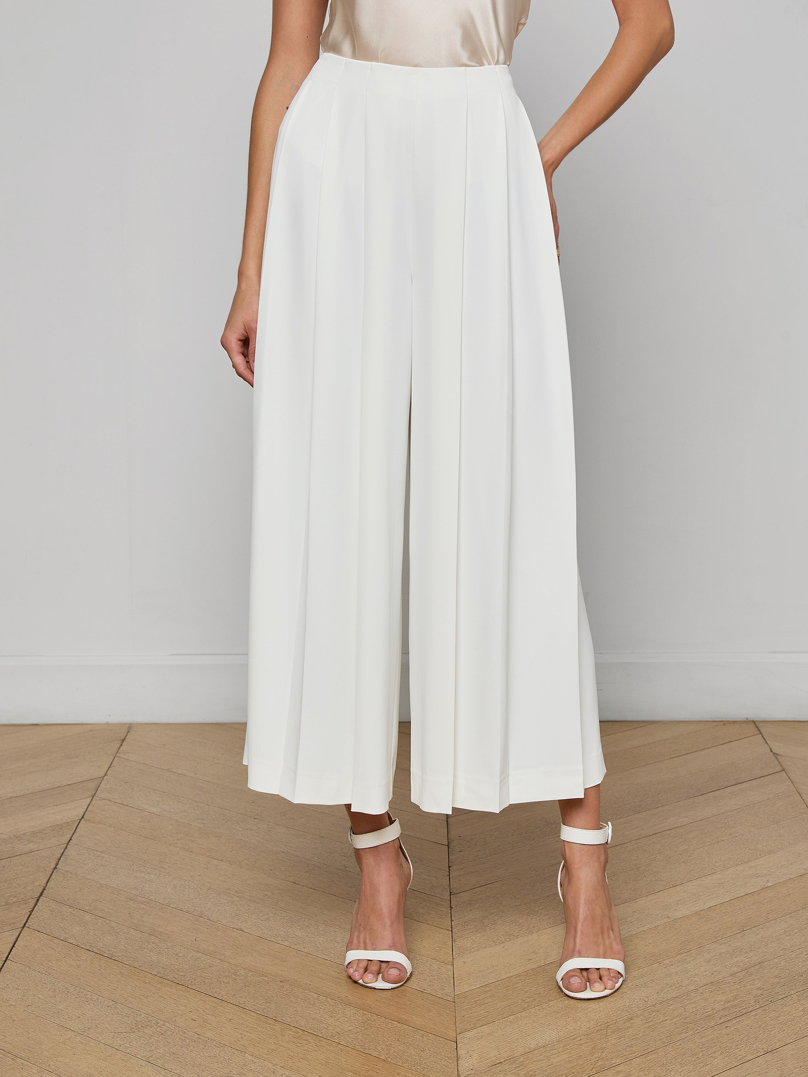 A woman stands against a plain light background wearing the Novalynn Cropped Pleated Pant in white—a high-rise, wide-leg culotte style—paired with a sleeveless beige top and white high-heeled sandals.