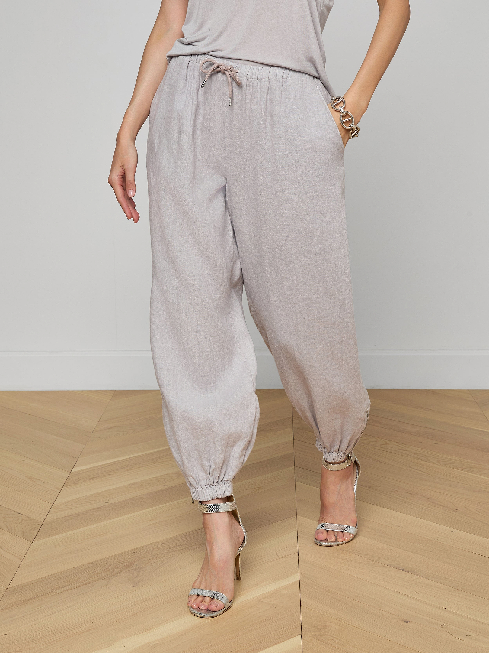 A person wearing light gray Mariko Linen Jogger pants, a matching top, and high-heeled sandals stands against a plain white background.