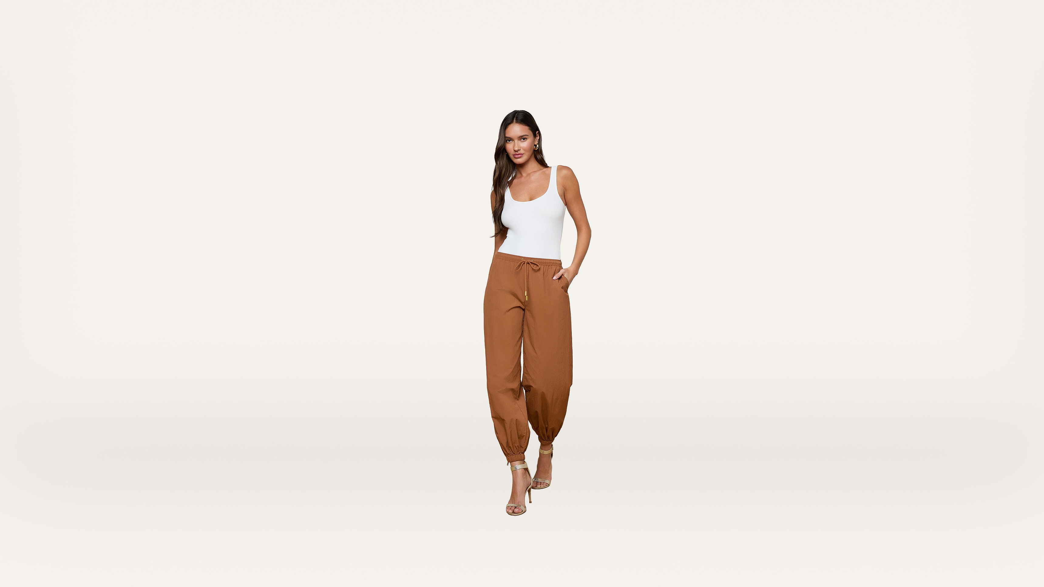 Woman wearing a white tank top and brown jogger pants with drawstring, standing and smiling with one hand in her pocket, on a plain white background.
