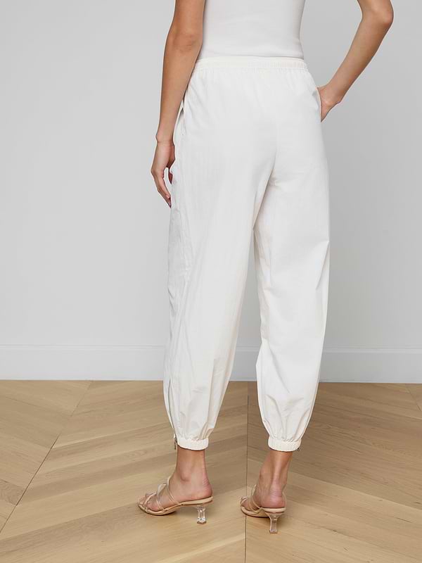 Someone is standing against a plain, light background, shown from the waist down and viewed from behind. They are wearing the Mariko Jogger—modern jogger-style pants in ivory crafted from water-repellent nylon—with heeled sandals.