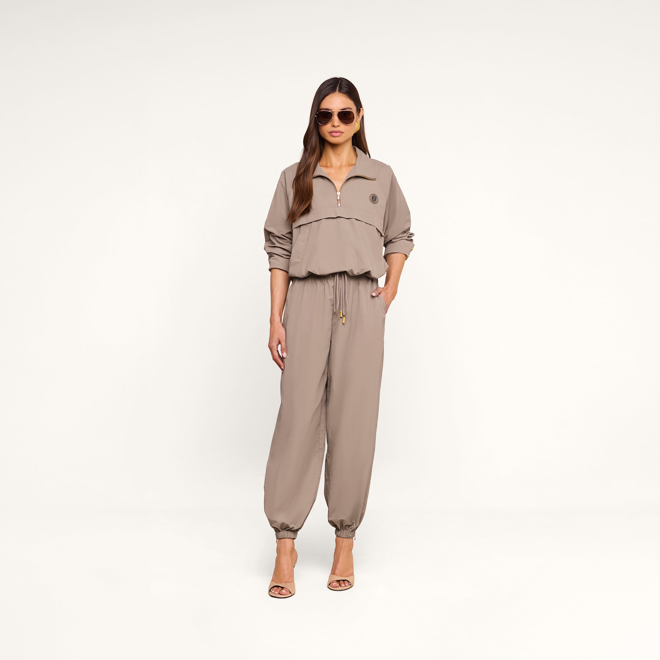 A woman wearing sunglasses and a light cardamom brown, long-sleeve zip-up top with matching Mariko Jogger pants crafted from water repellent nylon and heeled sandals stands against a plain white background.