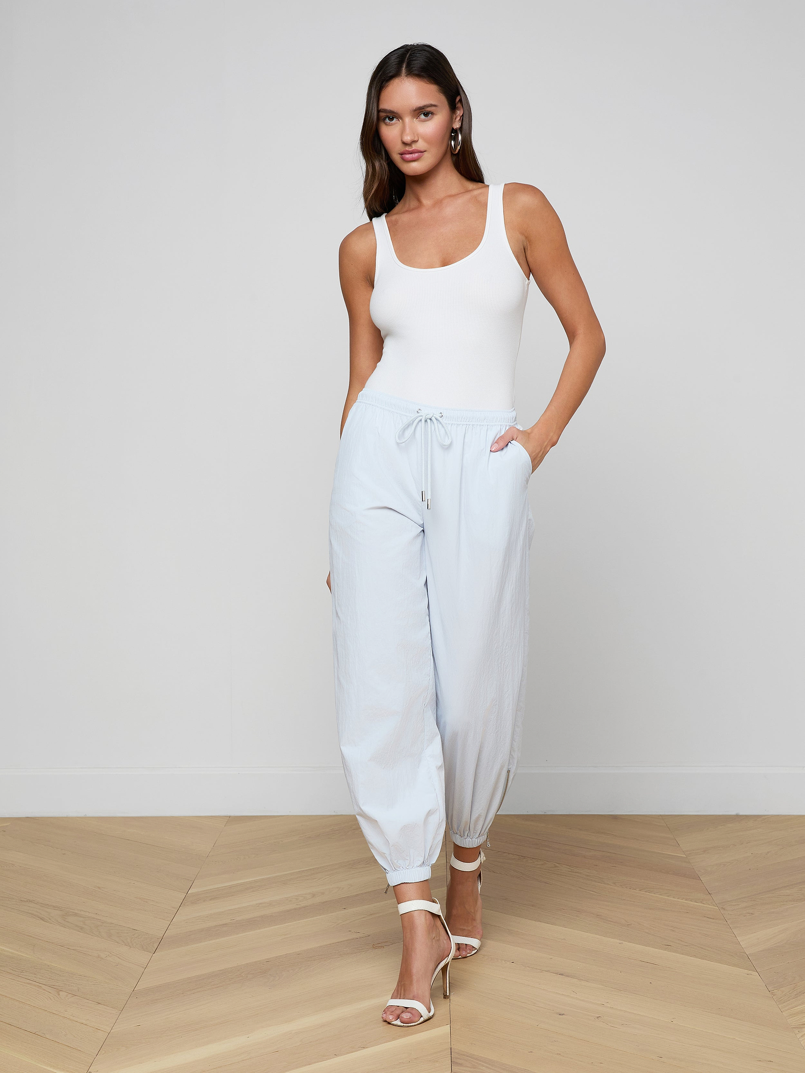 A woman stands against a plain white background wearing a white tank top, the Mariko Jogger in light blue, and white high-heeled sandals, with her hands in her pockets.
