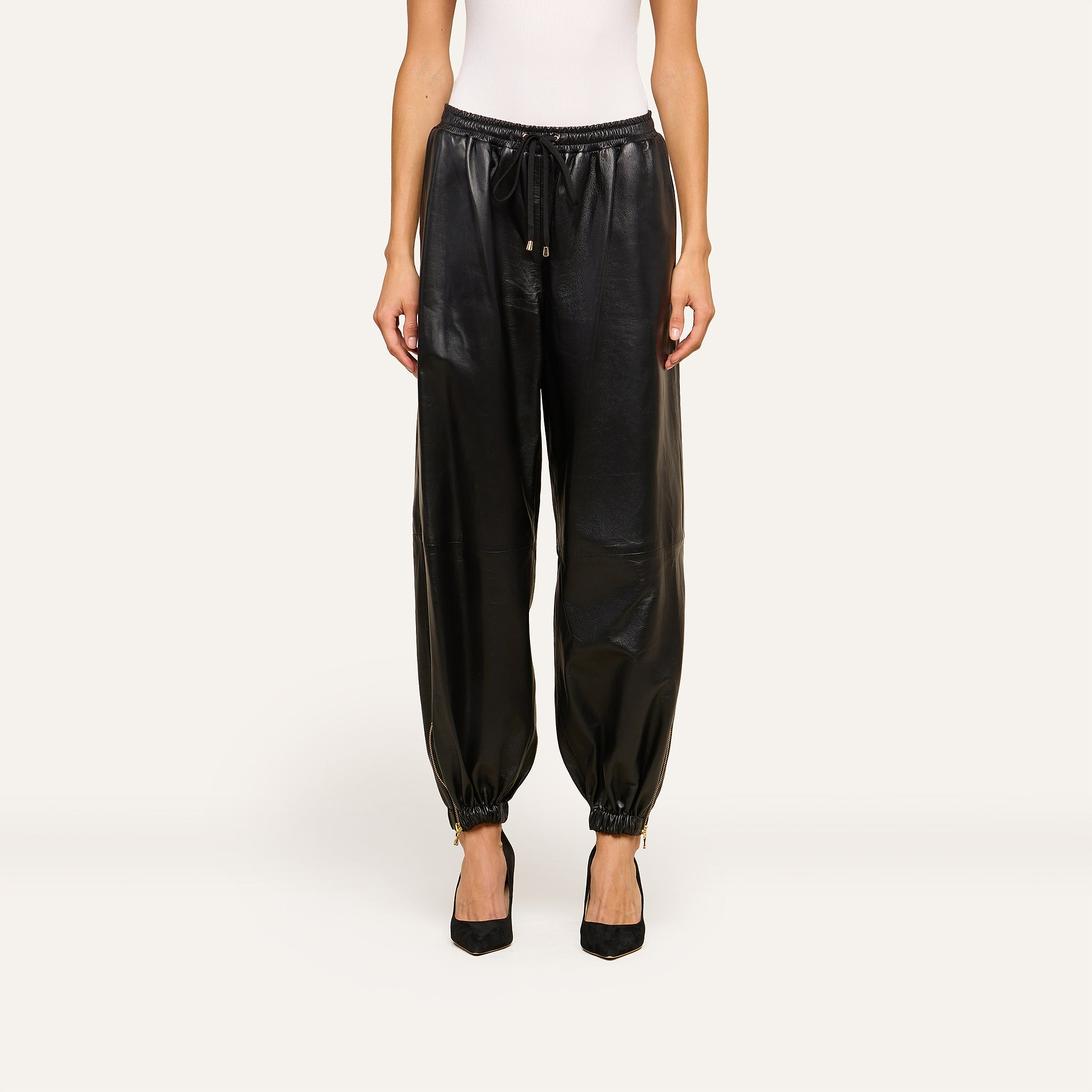 Wearing the Mariko Leather Jogger in black lambskin—with a drawstring waist and elasticated ankles—a person pairs the joggers with a white sleeveless top and black pointed-toe heels, standing against a light background.