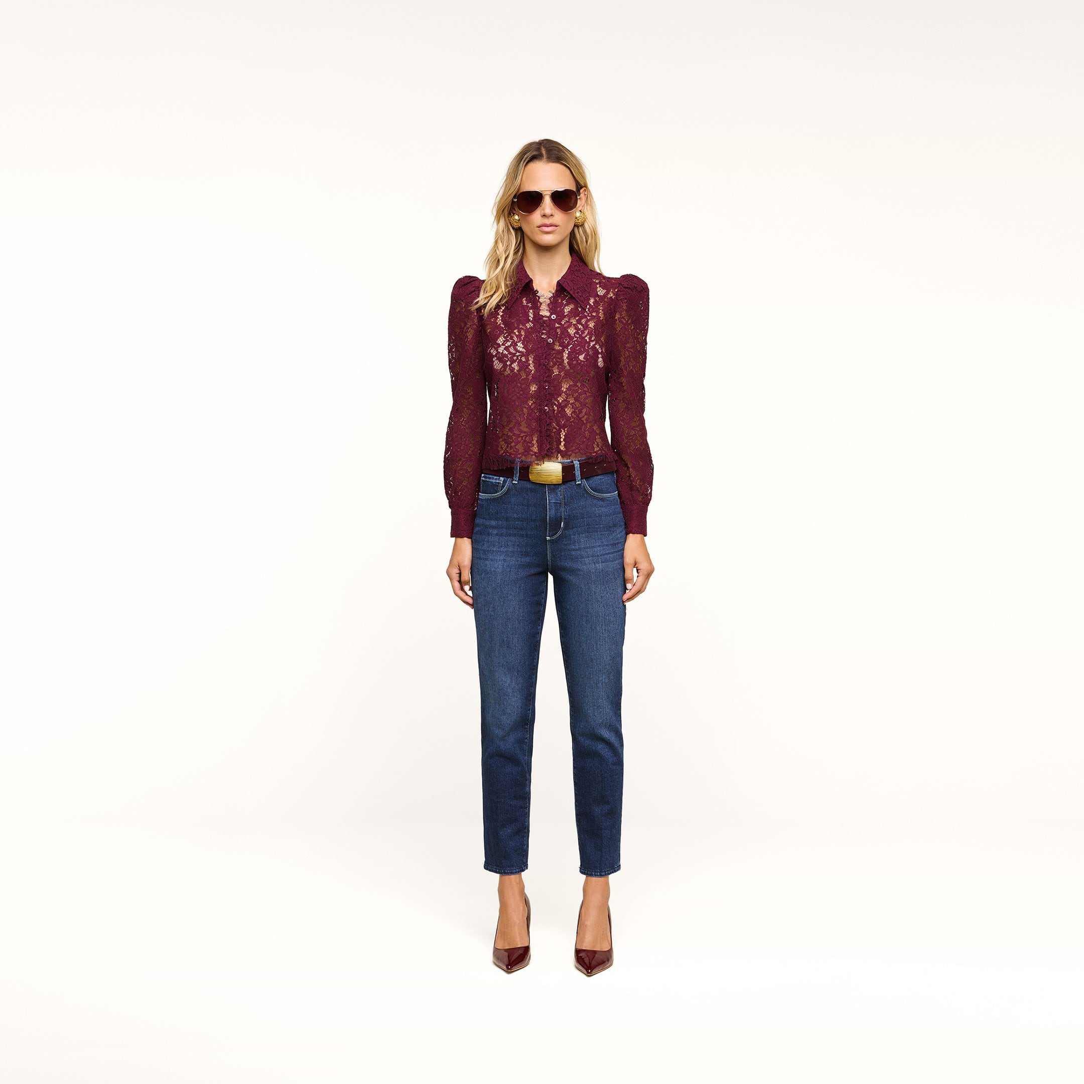A woman stands against a plain background wearing sunglasses, a burgundy lace blouse, and the Celestia Slim-Leg Jean in classic stretch denim with an ultra high rise fit. She completes her look with a brown belt and burgundy heels.