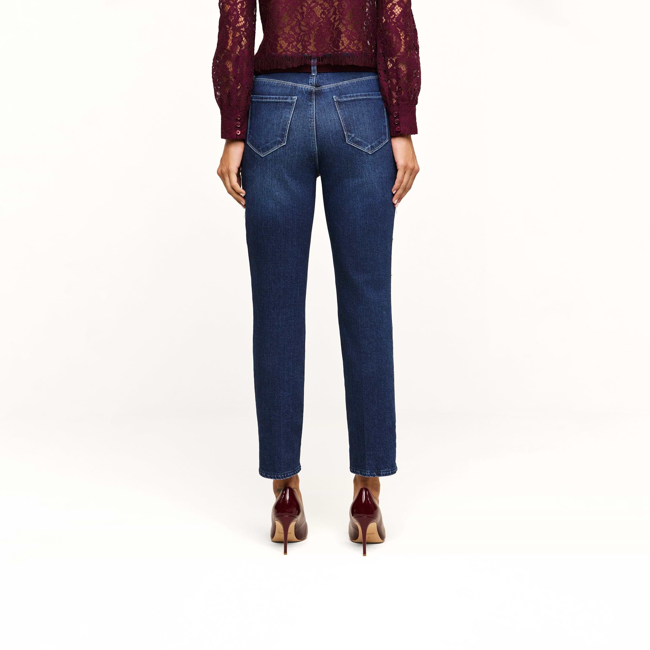 A person stands with their back to the camera, wearing a burgundy long-sleeve lace top, Celestia Slim-Leg Jean in a dark indigo wash stretch denim, and burgundy high-heeled shoes against a plain white background.