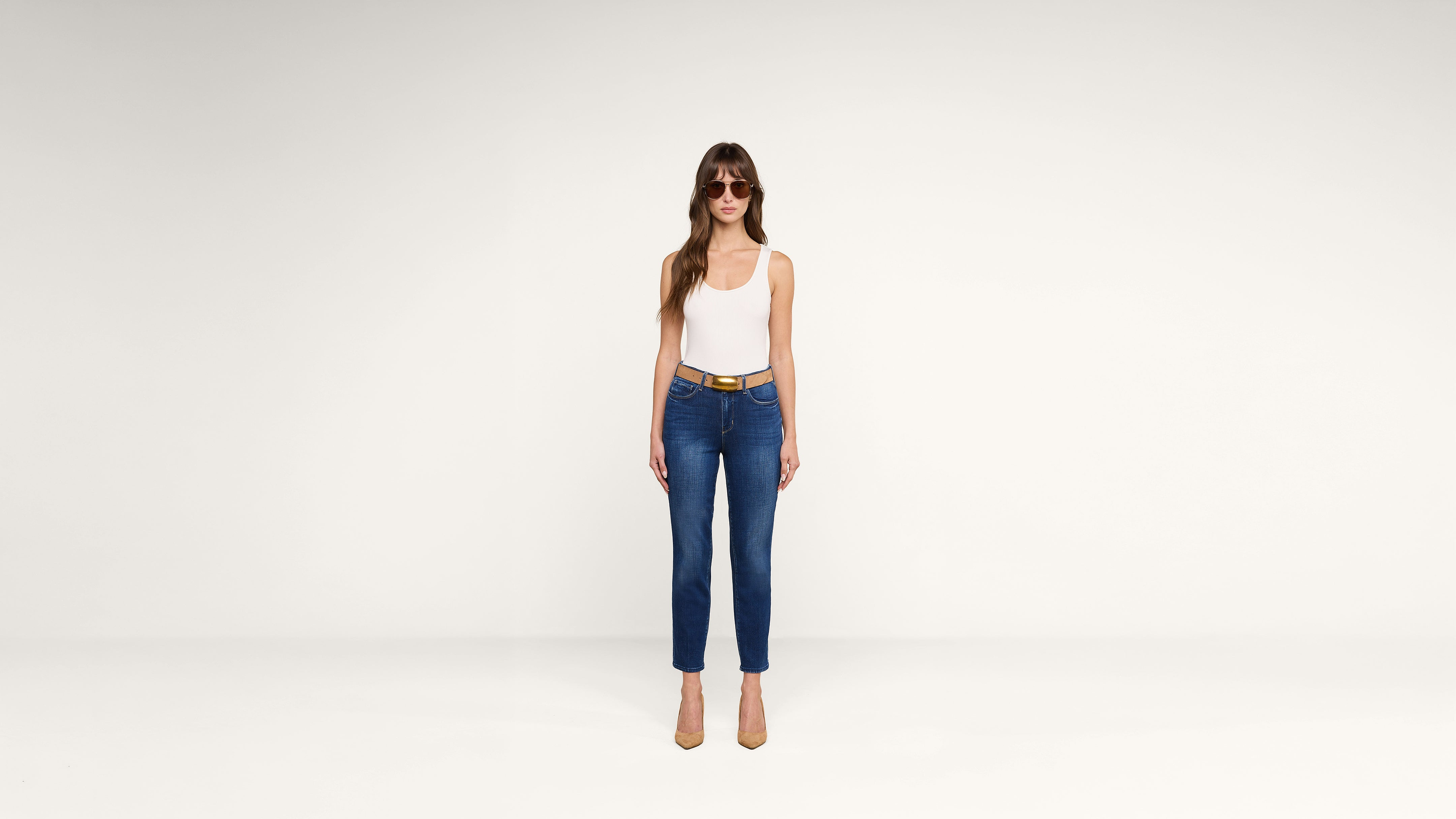 A woman with long hair wears sunglasses, a white tank top, blue jeans with a belt, and tan heels, standing against a plain white background.