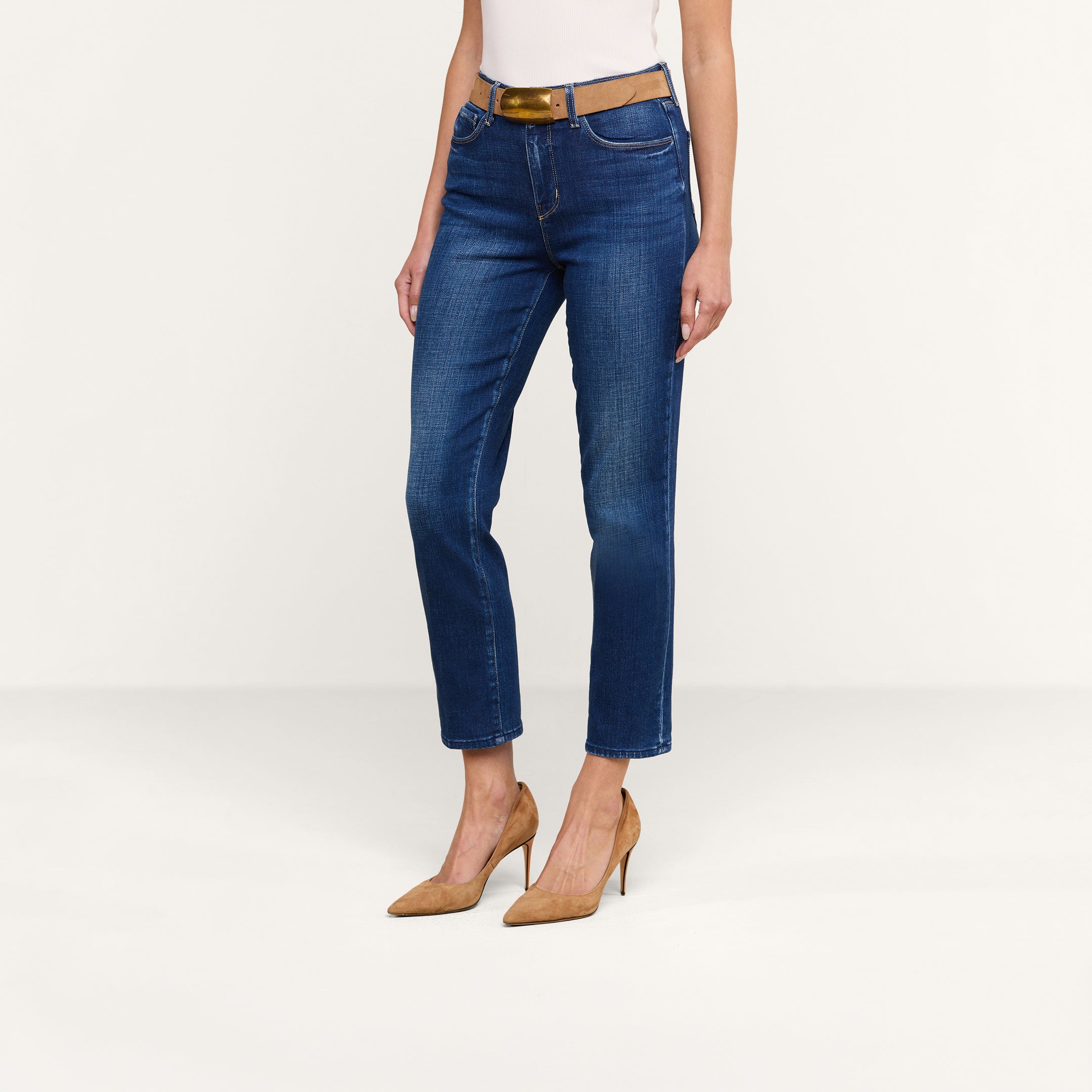Eine Person, die eine blaue „Celestia Slim-Leg“-Jeans, einen hellbraunen Gürtel, ein weißes Oberteil und hellbraune Schuhe mit hohen Absätzen trägt, steht vor einem beigen Hintergrund.