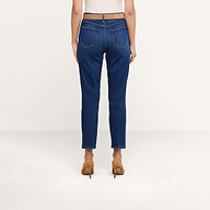 Eine Person steht mit dem Rücken zur Kamera und trägt ein weißes Oberteil sowie eine blaue „Celestia Slim-Leg“-Jeans aus Stretch-Denim, dazu hellbraune Schuhe mit hohen Absätzen. Der Hintergrund ist einfarbig beige.