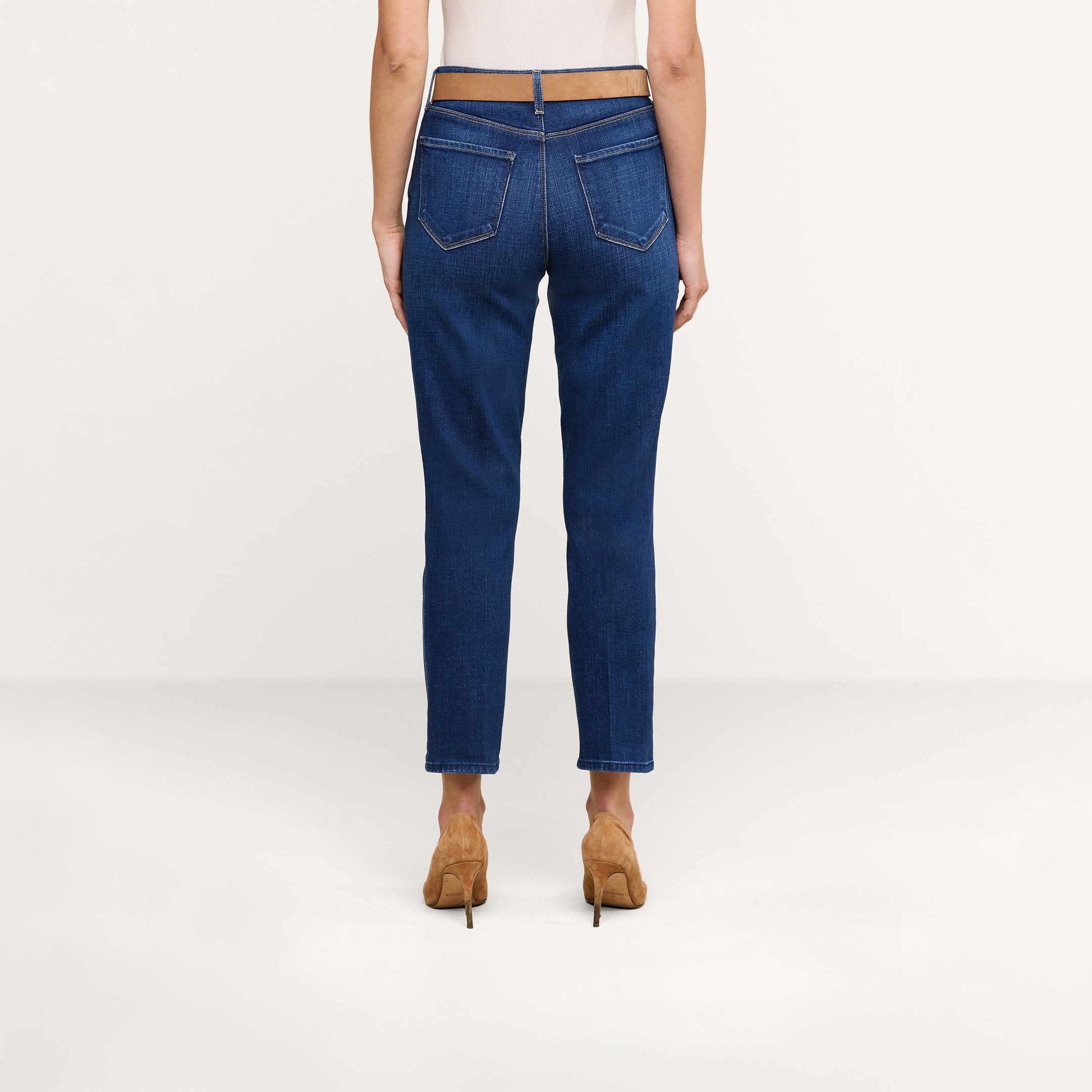 Eine Person steht mit dem Rücken zur Kamera und trägt ein weißes Oberteil sowie eine blaue „Celestia Slim-Leg“-Jeans aus Stretch-Denim, dazu hellbraune Schuhe mit hohen Absätzen. Der Hintergrund ist einfarbig beige.