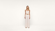 A woman with long blonde hair stands facing forward, wearing a sleeveless beige jumpsuit with wide legs and a tan belt, against a plain white background.